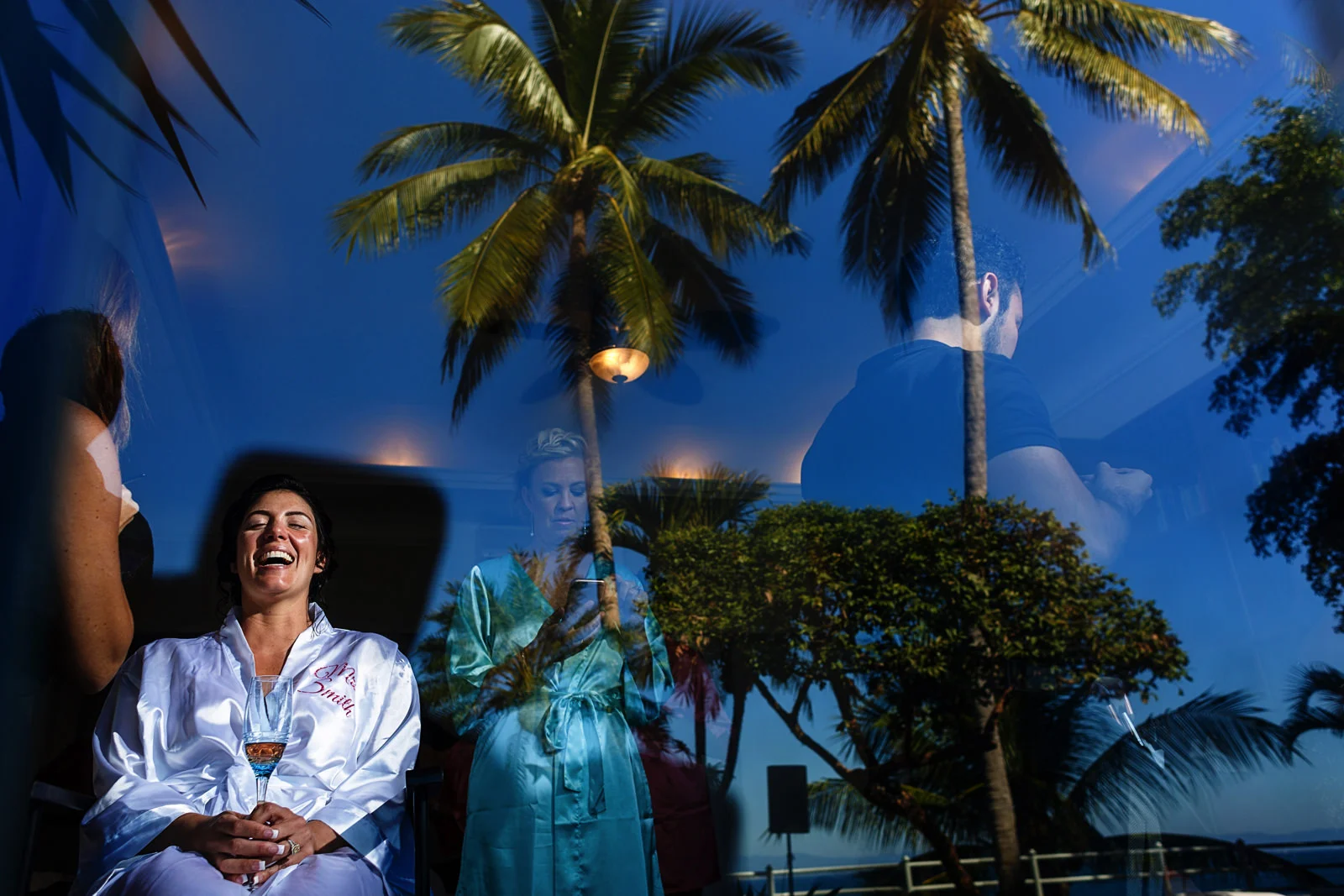 Bride laughing while she is having her make-up, through a window that also reflects the blue sky and palm trees