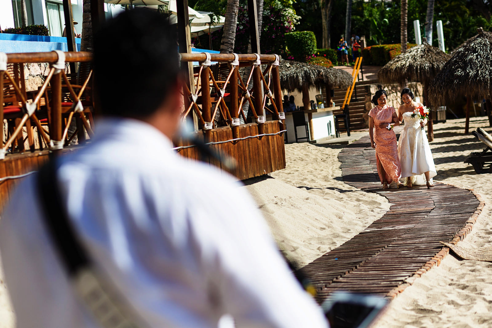 Bride and her mother walk on a wood-made path in the beach while a performer sings