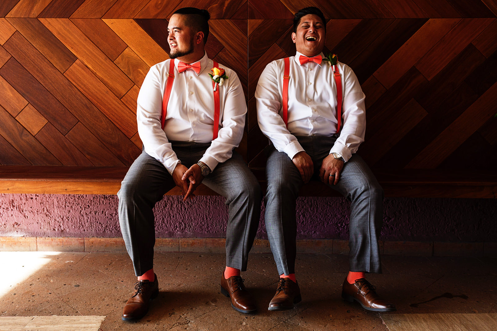 Groom and best man wearing matching outfits laugh sitting on a bench