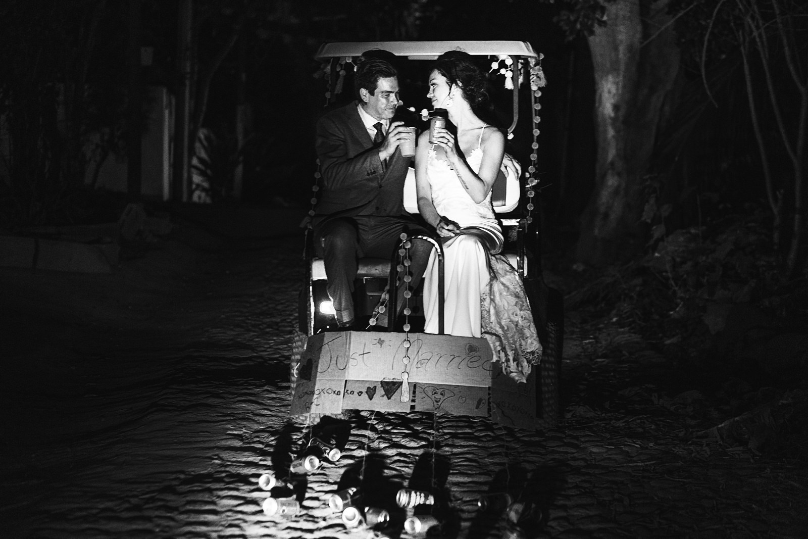 Bride and groom exiting the wedding ceremony on a golf cart that has a “just married” sign, while they cheer with beers