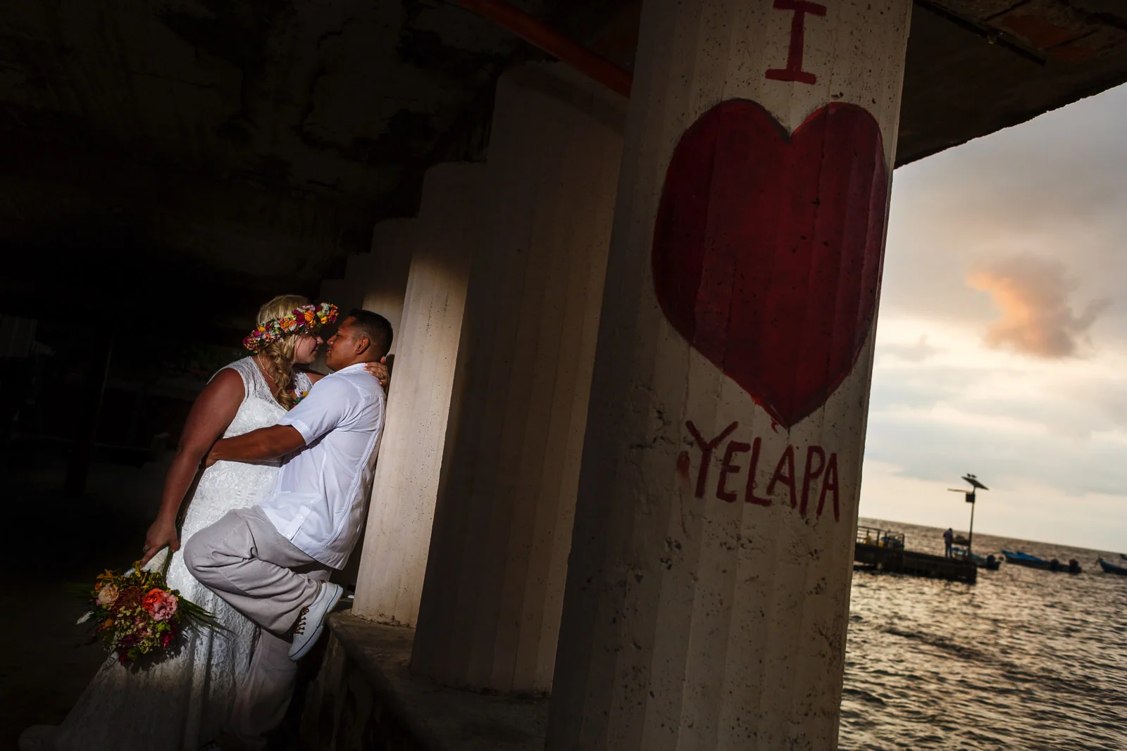 Bride and groom leaning against a column with "I <3 Yelapa" painted on, and the sun setting in the background.