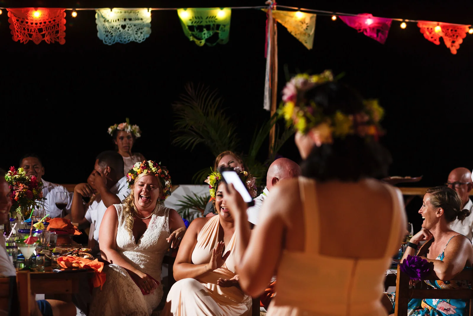 Bride and the rest of the wedding guests laughing during the bridesmaid’s speech that she is reading from her phone.