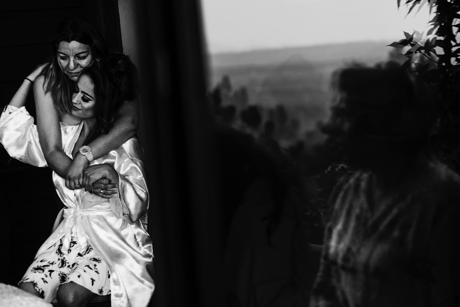 A friend of the bride hugs her from behind while she is on the getting ready at her bedroom, a reflection on the window shows the valley of Atemajac.