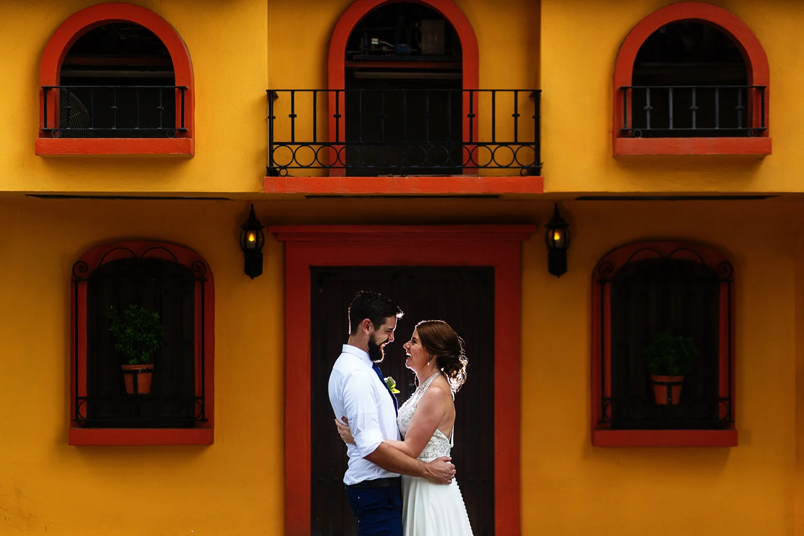 Bride and groom laughing facing each other outside a Mexican styled facade.