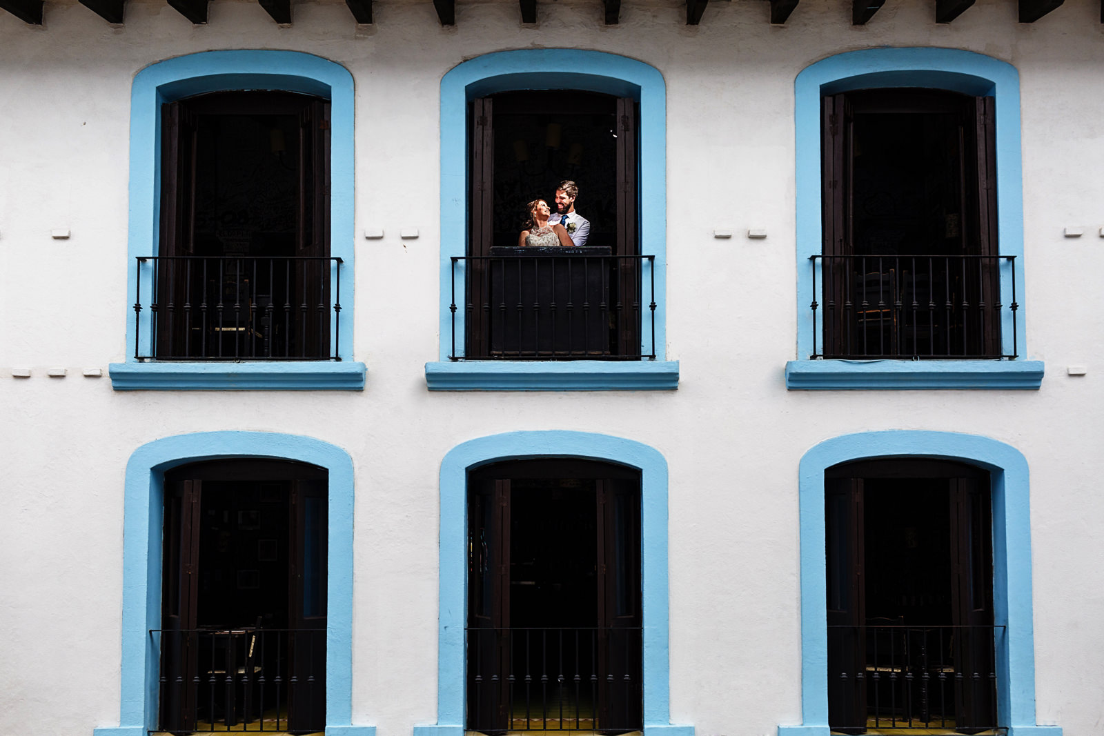 Groom and bride portrait, the couple is framed on a blue window with a white facade.