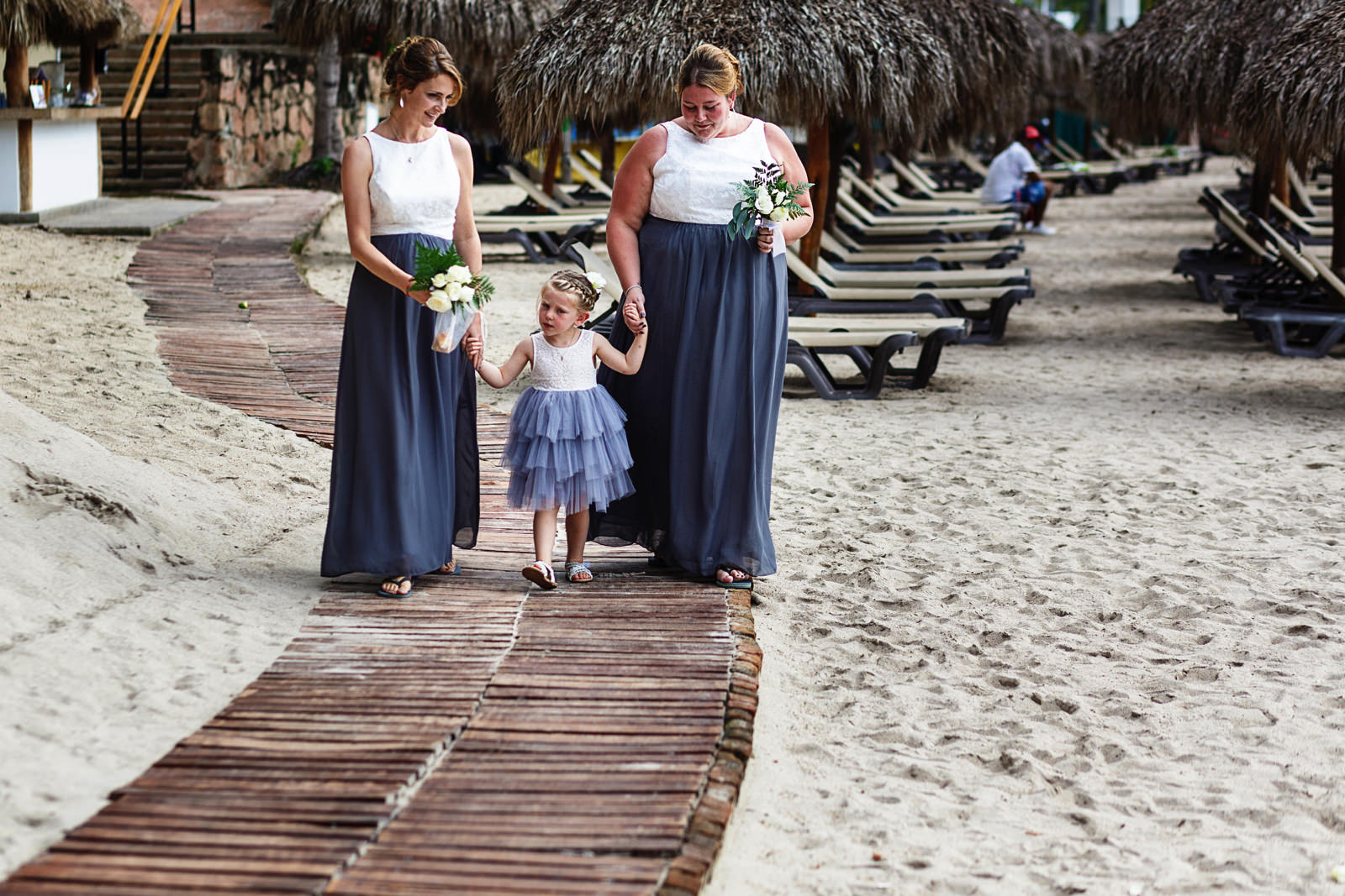 Bridesmaids and flower girl walking down a wood path on the beach for the wedding ceremony to start.