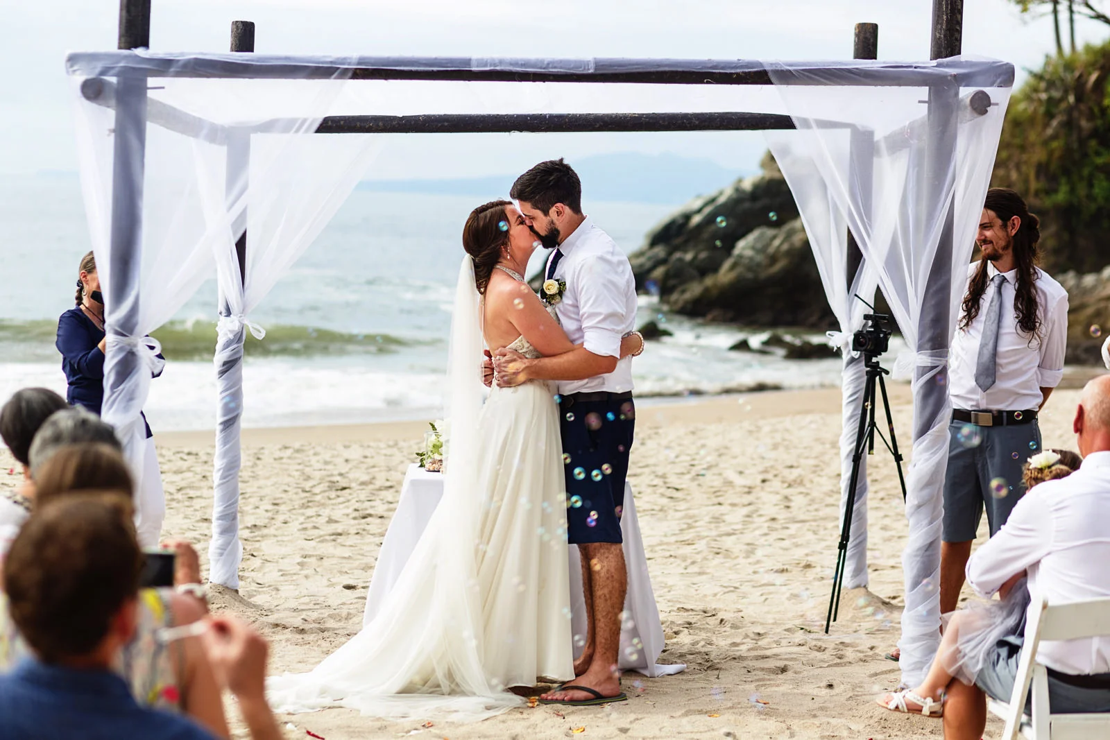 Bride and groom first kiss while the wedding guests blow bubbles at the end of their beach wedding ceremony.