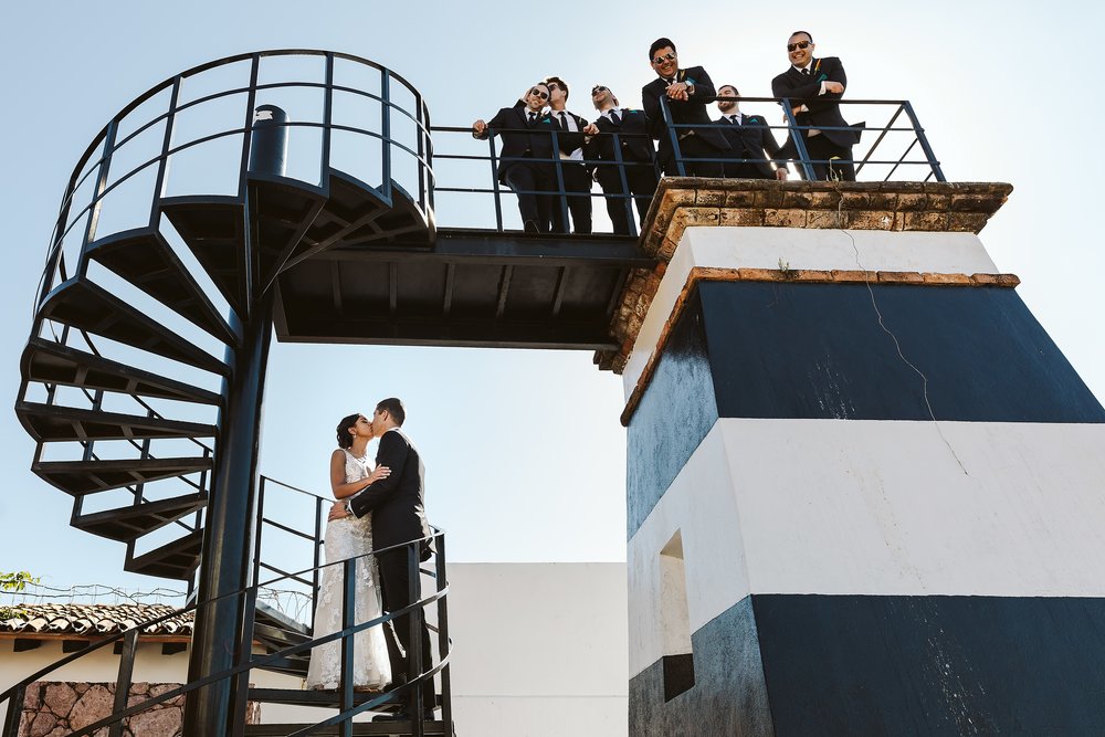 Bride and groom kiss on a spiral staircase while groomsmen watch and kiss as well