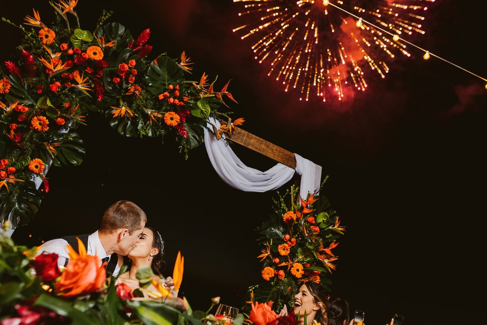 Groom and bride kiss as fireworks explode against the dark night sky