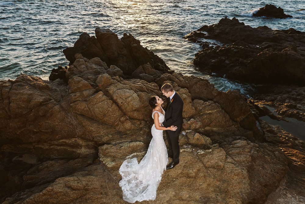 Bride and groom smile at each other standing on rocks near the ocean in Puerto Vallarta at sunset