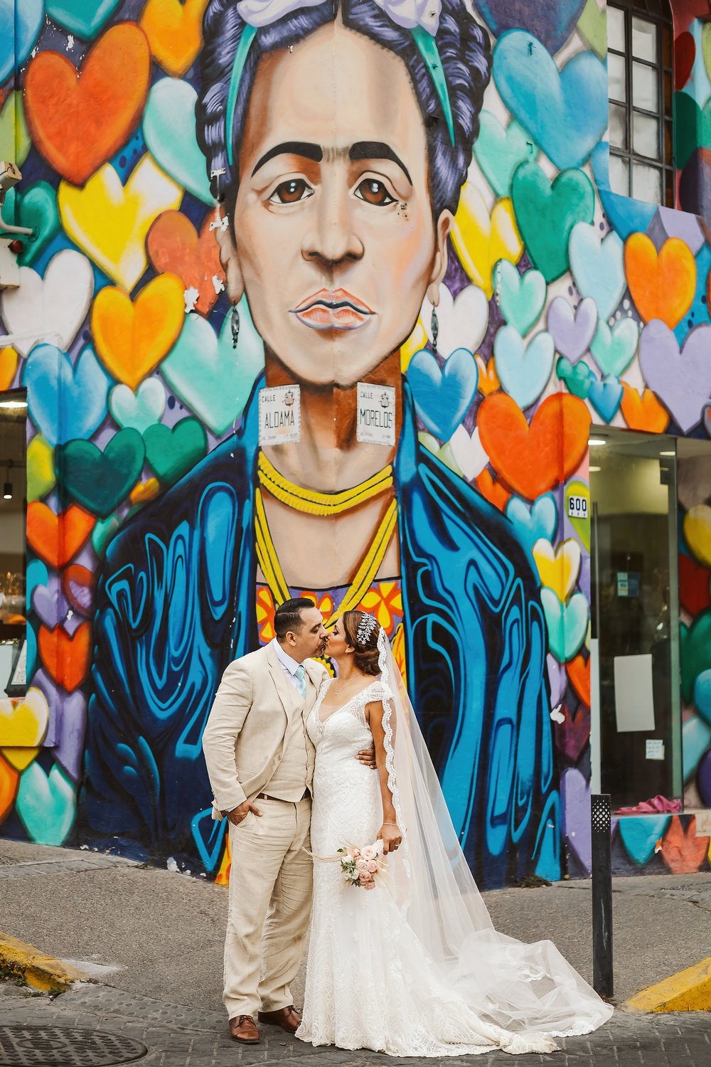 Bride and groom kiss in front of a big mural wall with a Frida Kahlo on it