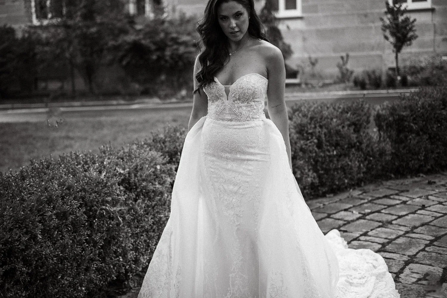 Montreal wedding photographer photographed a bride in a wedding dress standing outdoors on a cobblestone path surrounded by bushes, with a building and trees in the background.