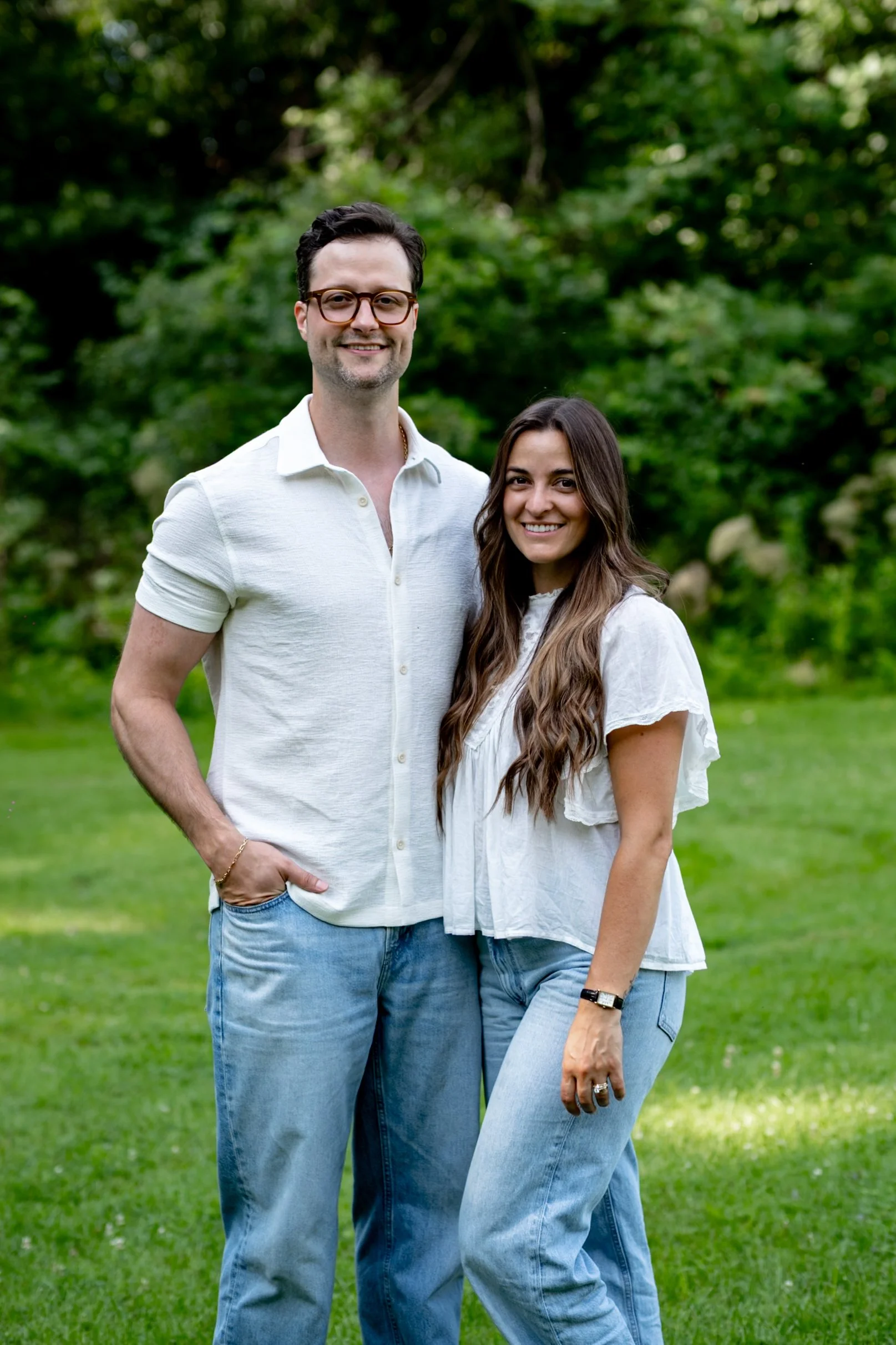 A young couple smiling outdoors in a green park. The man is wearing glasses, a white short-sleeved shirt, and jeans. The woman has long wavy brown hair, is wearing a white blouse, and light blue jeans. They are standing close together, with trees and