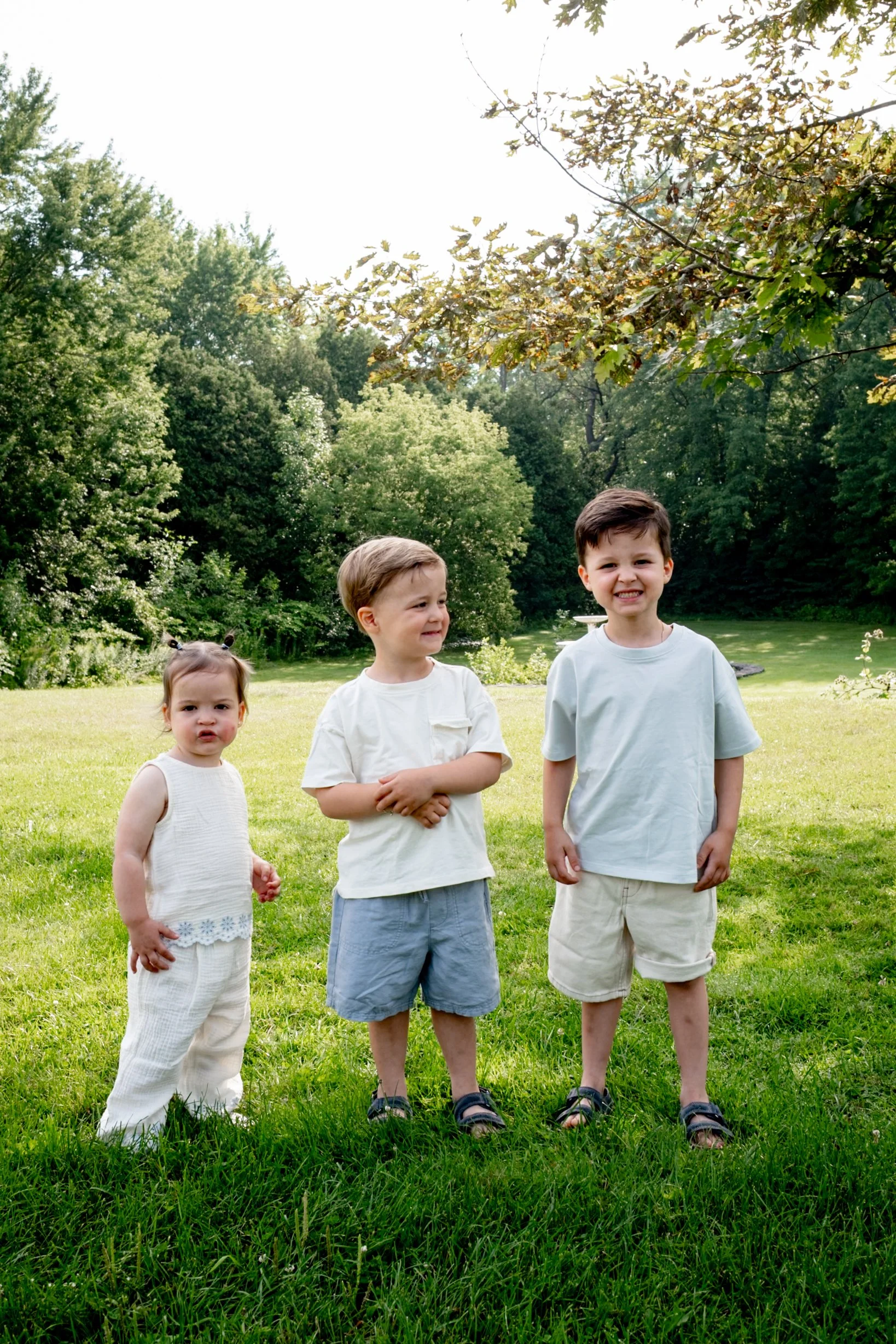 Three children standing on a grassy field with trees in the background, smiling and looking at the camera.
