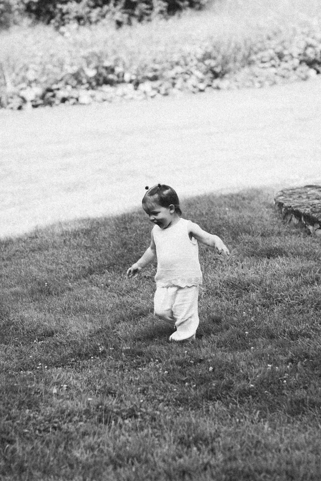 A young child with short hair and pigtails, wearing a sleeveless top and loose pants, standing and playing on a grassy lawn with a joyful expression.