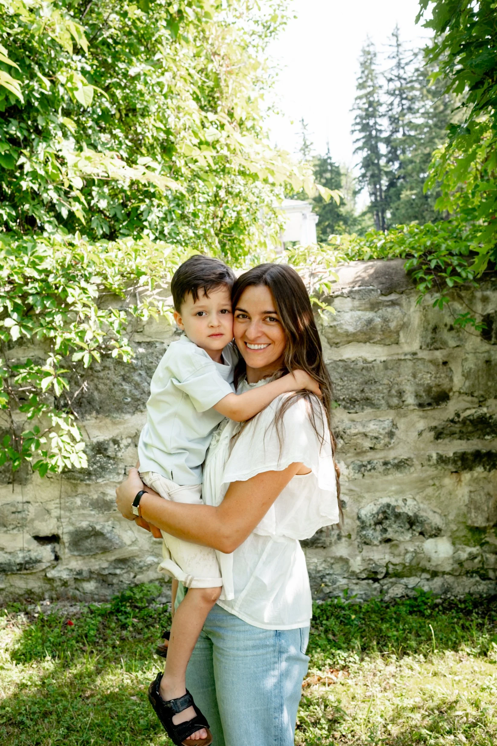 A smiling woman holding a young boy in an outdoor garden with lush green foliage and a stone wall in the background.