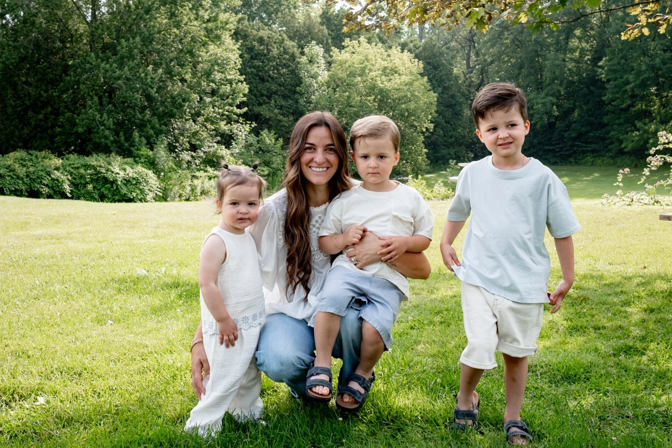 A woman with long brown hair smiling and sitting on the grass with three young children on a sunny day in a park with green trees in the background.