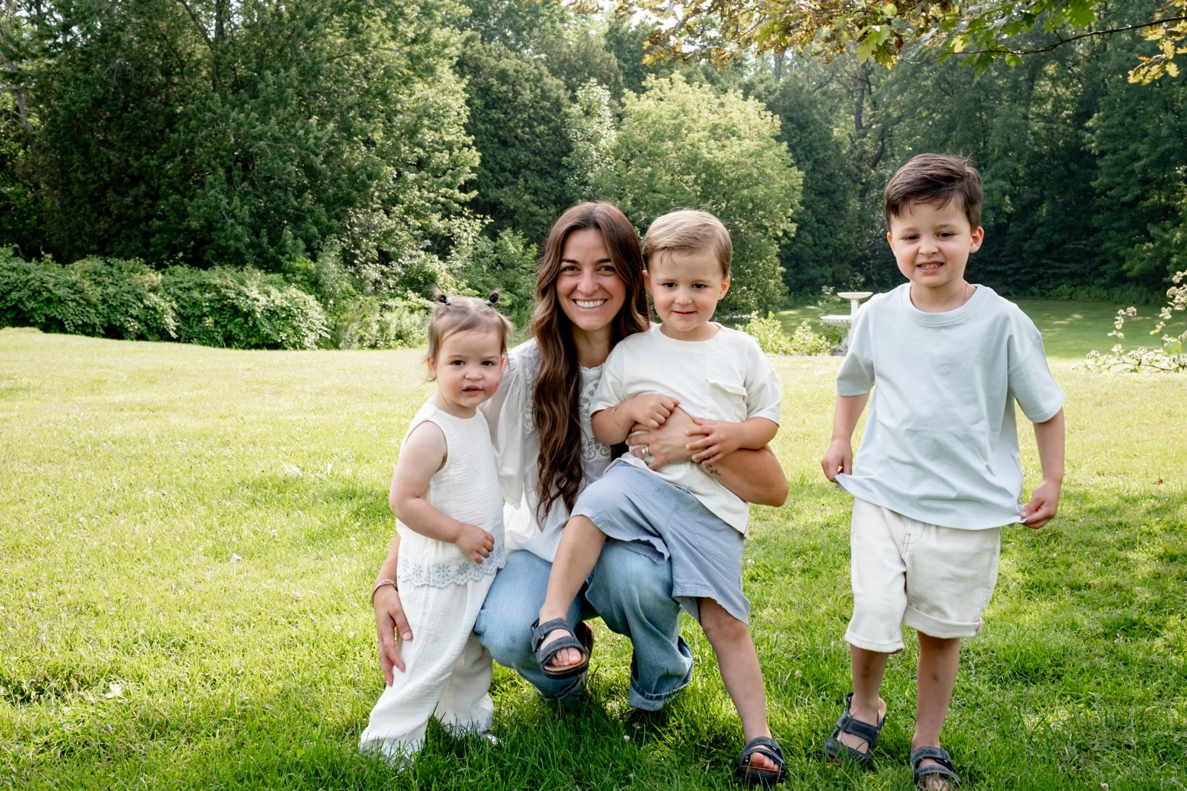 A smiling woman with long brown hair pose outdoors with four young children on a grassy lawn surrounded by trees.