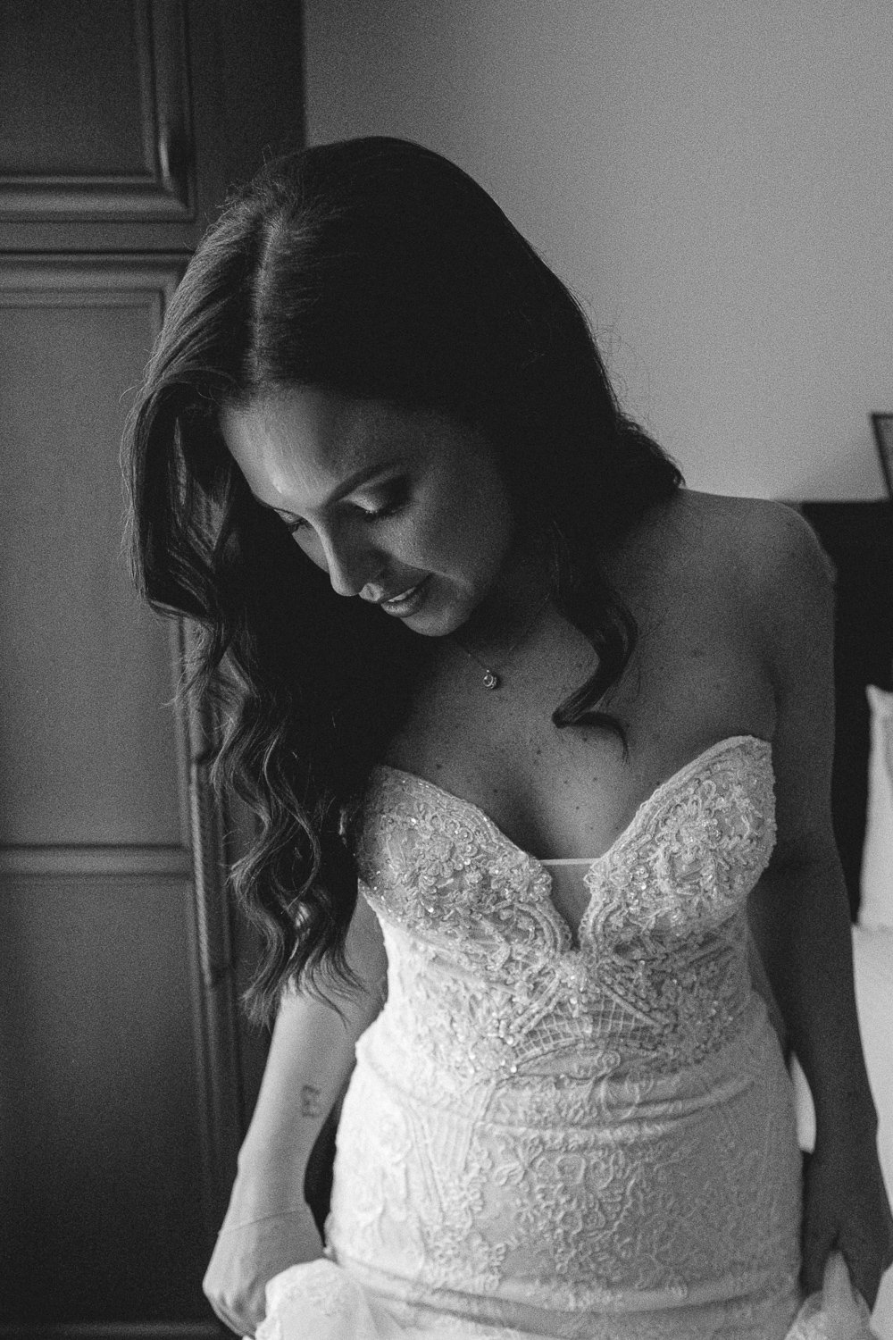 Black and white photo of a woman in a wedding dress, looking down with a smile, in a room with wardrobe and bed.