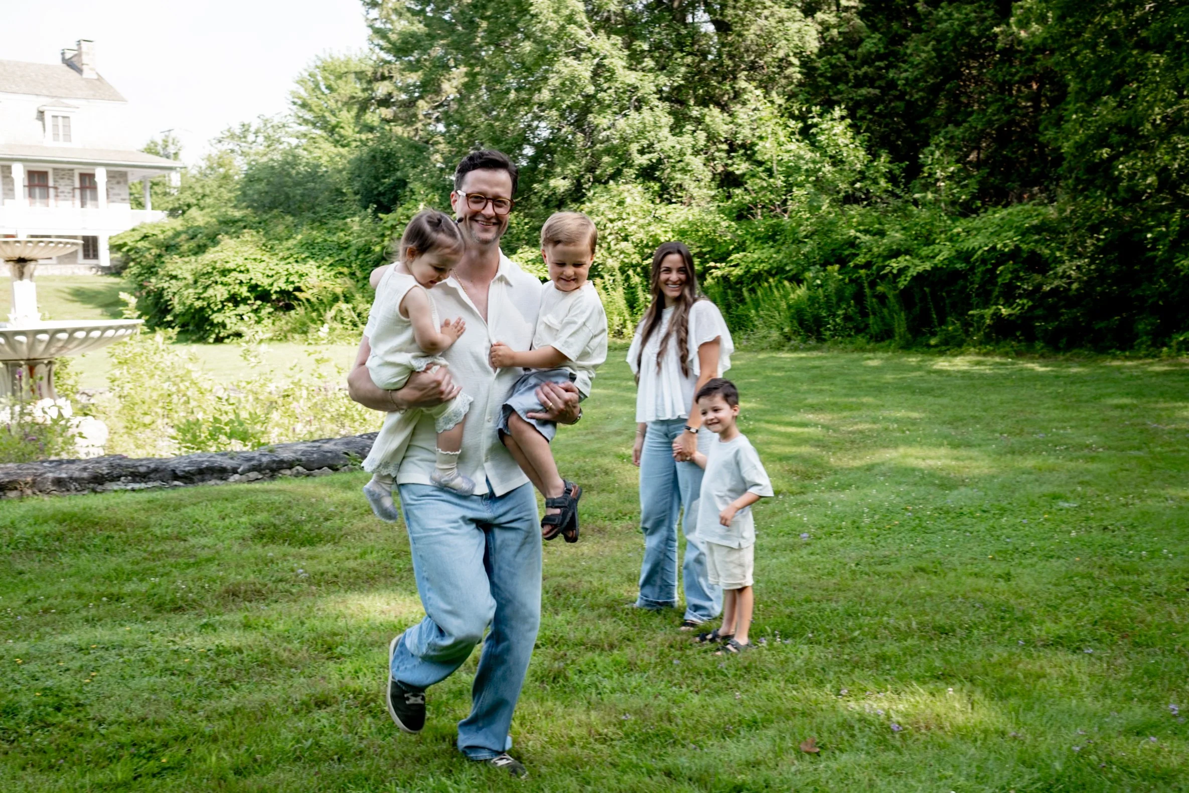 A family enjoying a day outdoors on a grassy lawn surrounded by trees. The father is carrying two children, and two other children and a woman are smiling nearby.