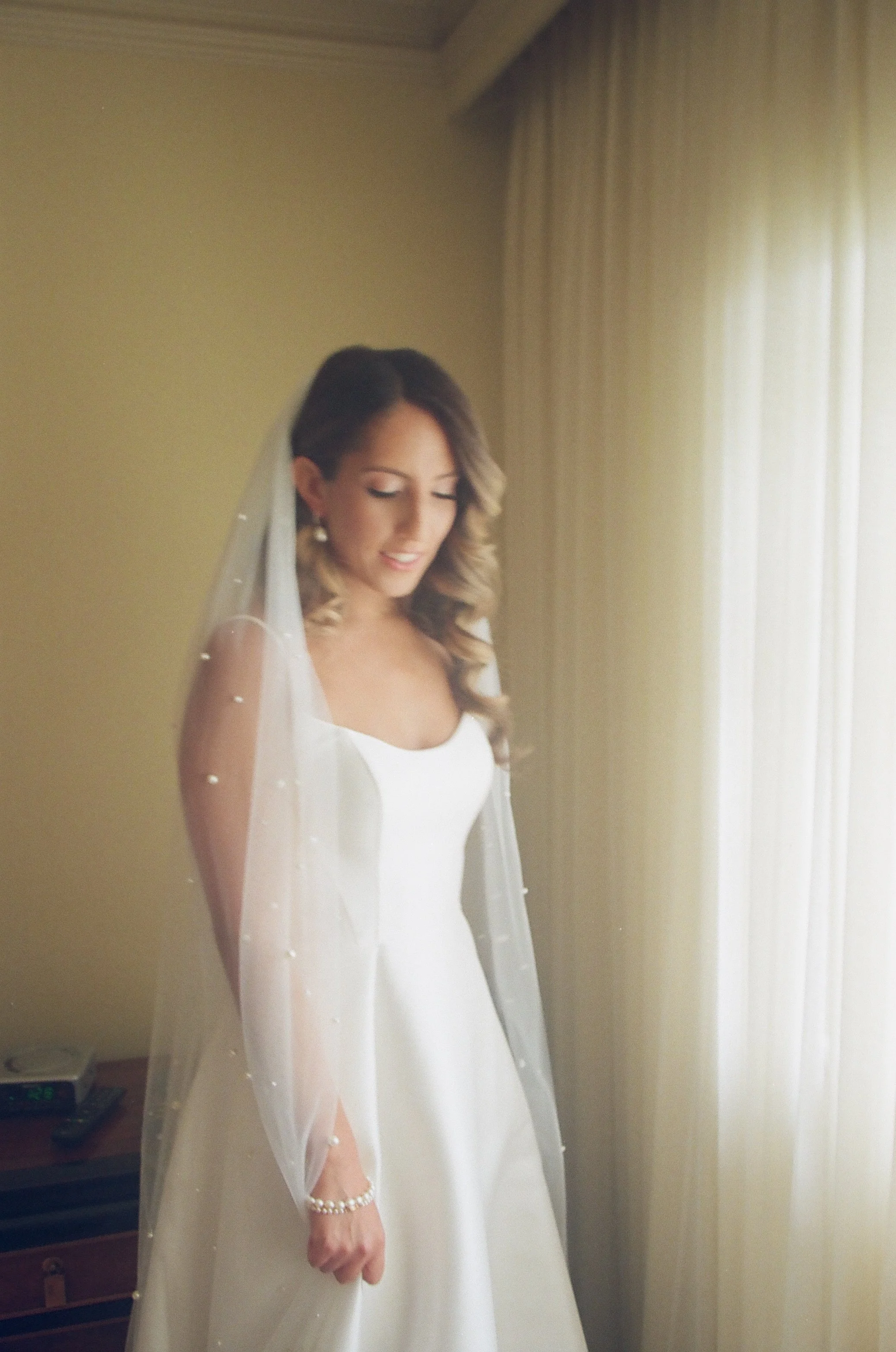 A bride standing indoors near a window with sheer curtains, dressed in a white wedding gown with a sleeveless design, wearing earrings and a pearl bracelet, and a veil with small embellishments.
