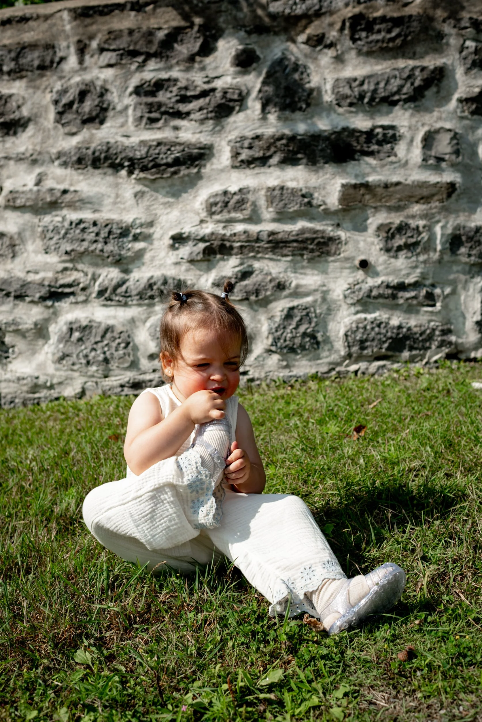 A young girl sitting on the grass in front of a stone wall, wearing a white dress and white sneakers.