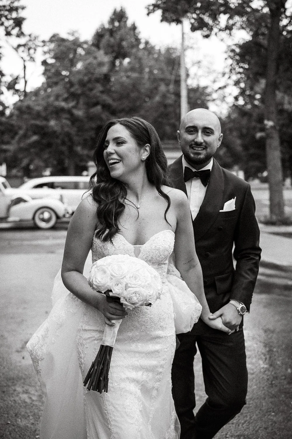 A black and white photo of a bride and groom walking outdoors, with trees and vintage cars in the background. The bride is holding a bouquet of roses and is smiling, while the groom is dressed in a tuxedo with a bow tie, smiling and looking at the ca