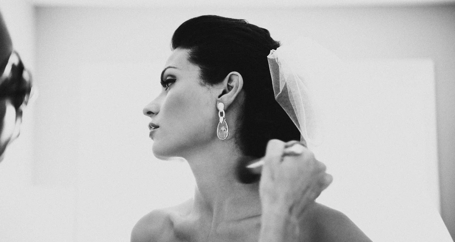 A woman with dark hair and fair skin getting ready, possibly for a wedding, as a stylist adjusts her hair. She wears large, dangling earrings and a sheer veil, with her face turned in profile against a plain background.