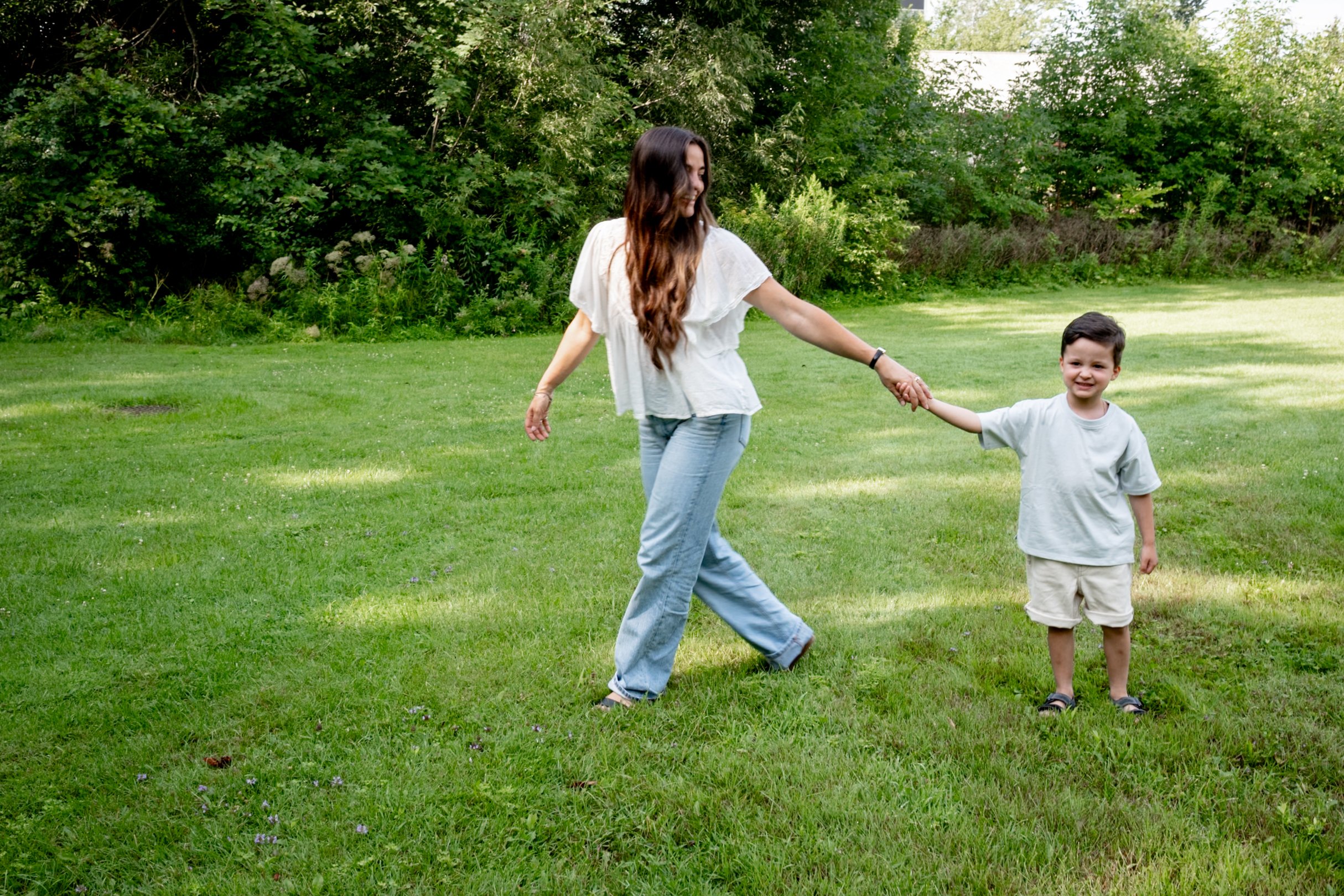 A woman and a young boy holding hands and walking on a grassy field with trees in the background.