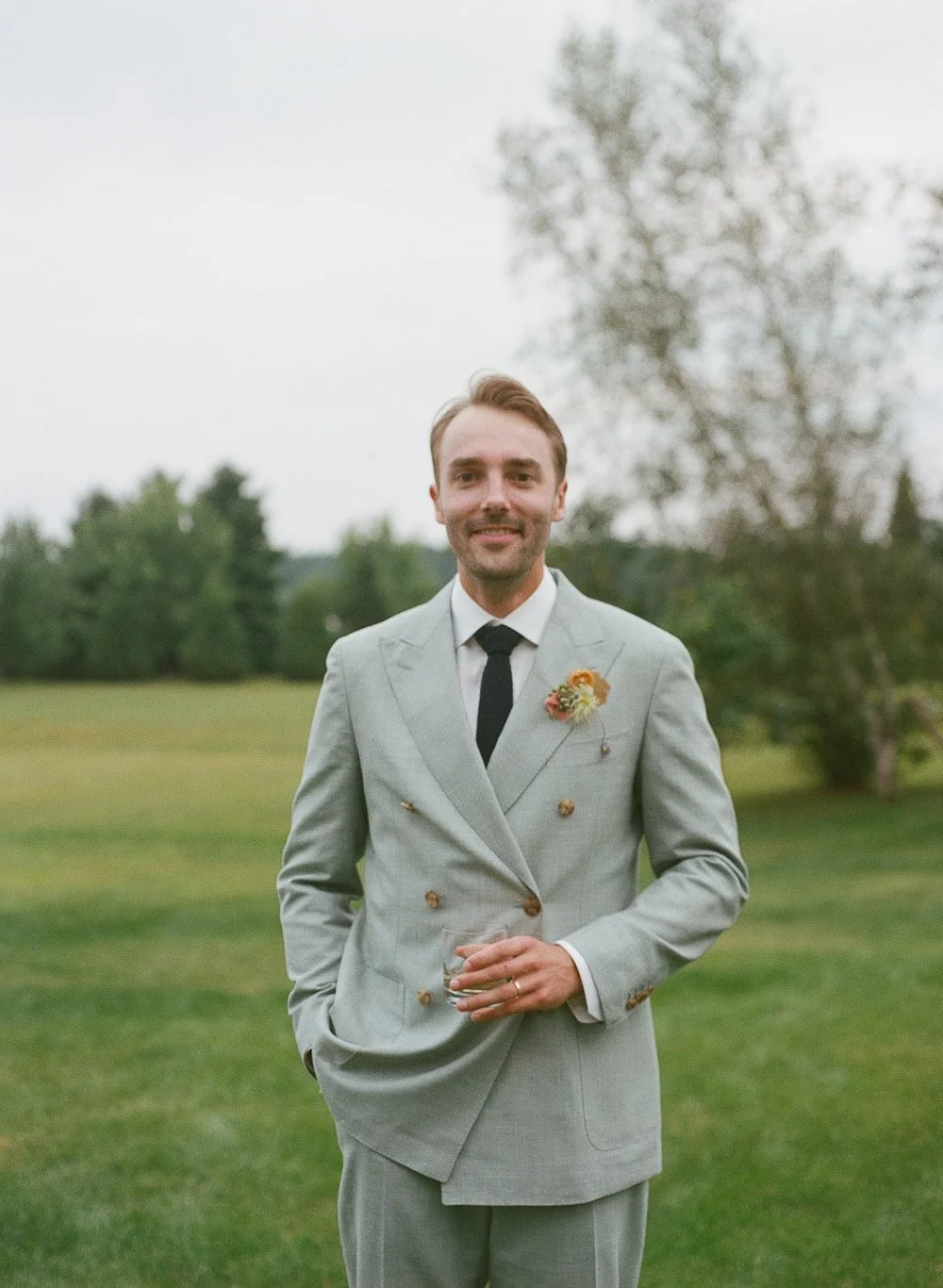 A man in a light gray suit with a boutonniere, black tie, and white shirt standing outdoors on a grassy field with trees in the background, holding a glass.