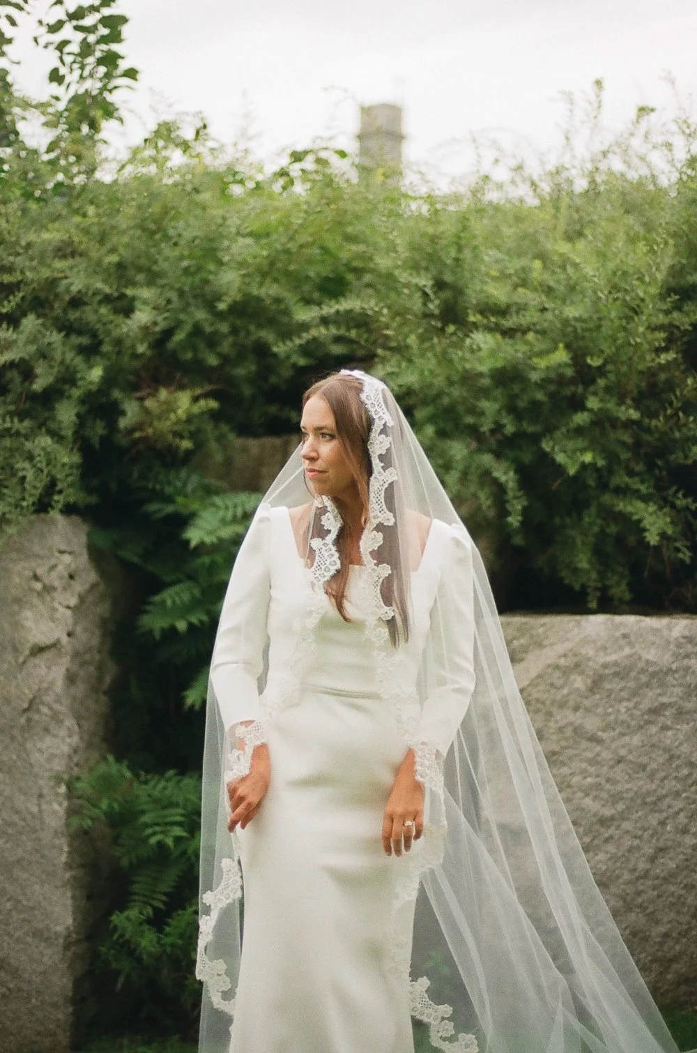 A woman in a white wedding dress and veil standing outdoors surrounded by green bushes and rocks.