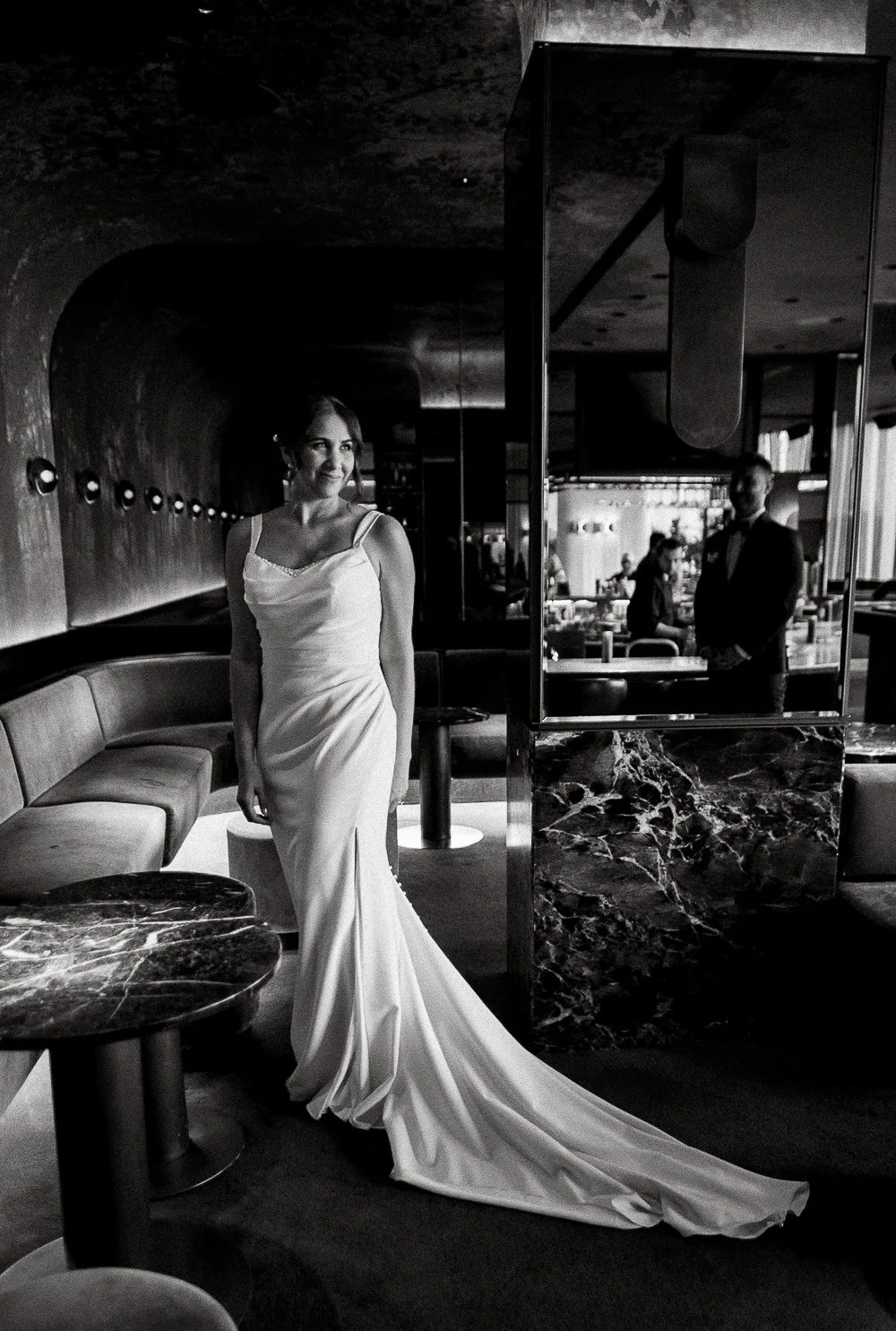 A woman in a white wedding dress standing in a stylish, modern restaurant or bar, with a man in formal attire visible through a mirror in the background.