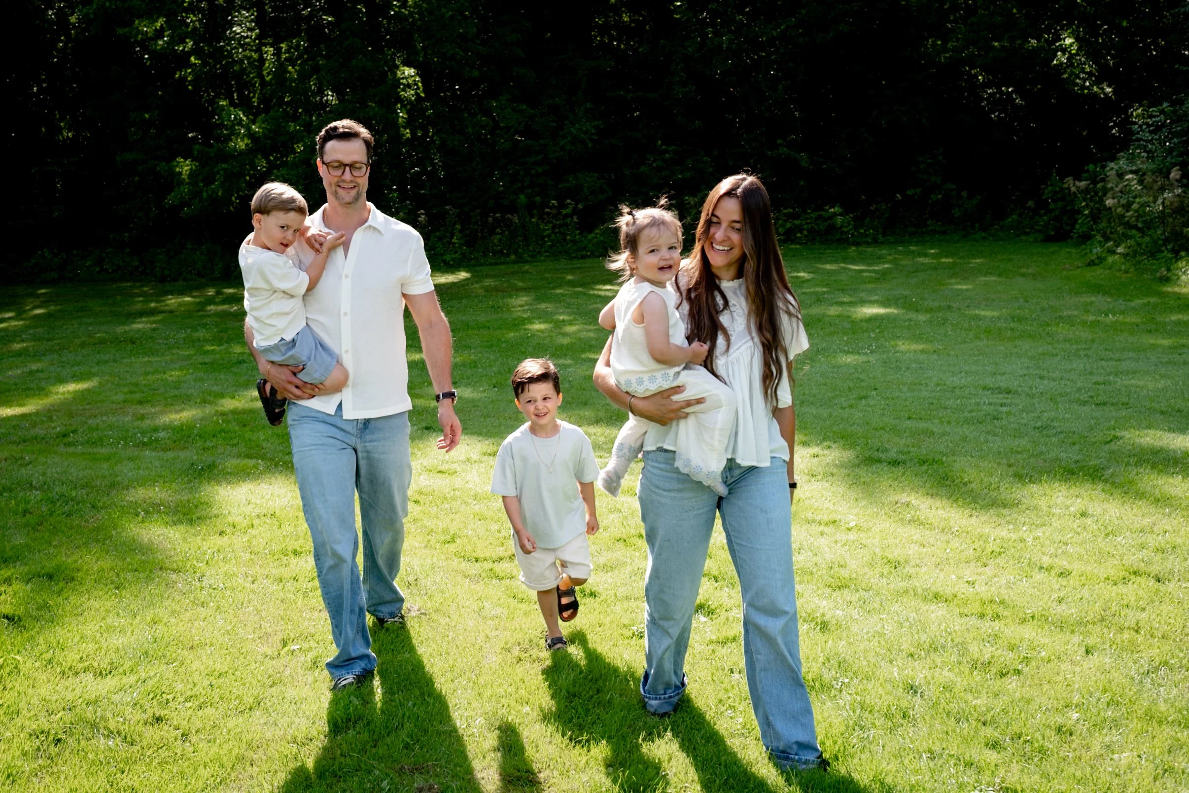 A family of five walking outdoors on a sunny day, with a green grassy area and trees in the background. The mother holds a young girl, and a man holds a young boy, while another boy walks beside them.