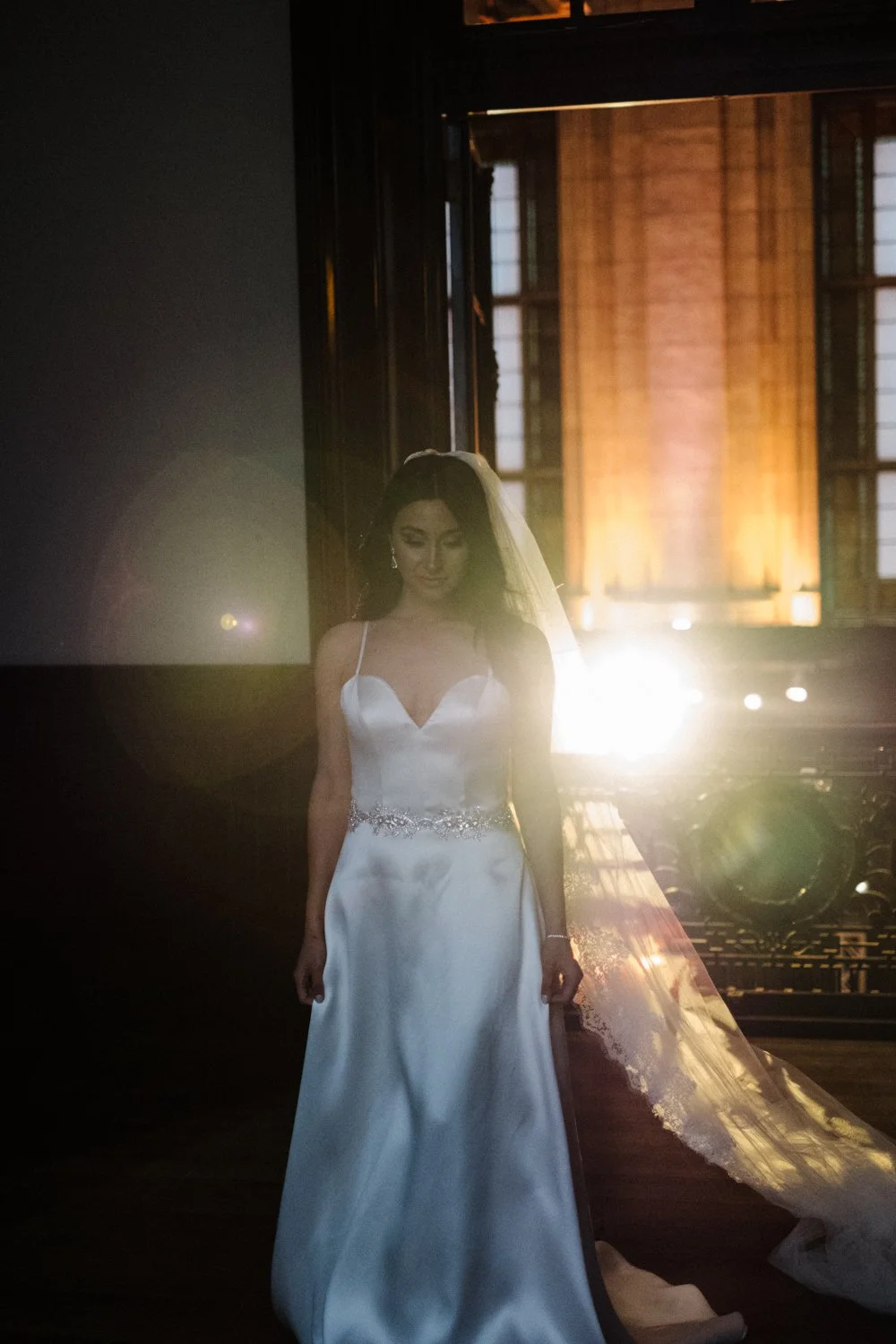 A bride in a white wedding dress with a veil, standing indoors with sunlight streaming in through large windows behind her.