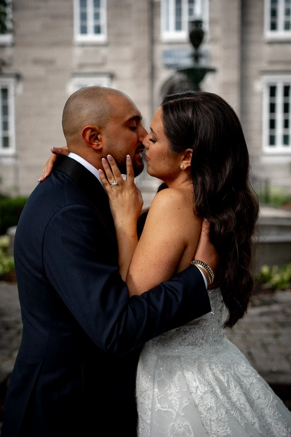 A newlywed couple sharing a close, intimate moment outdoors in front of a historic building, with the groom in a black suit and the bride in a white lace wedding gown.