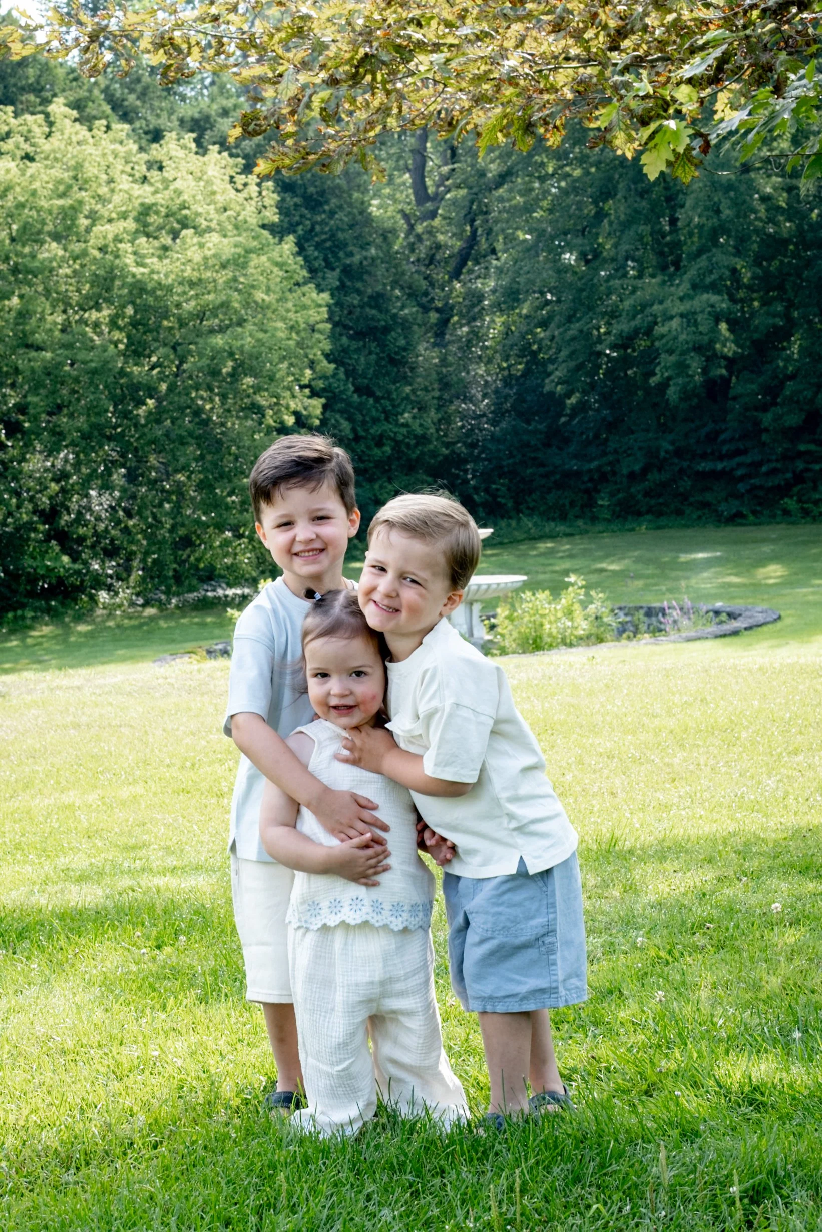 Four children, two boys and two girls, standing on a grassy field in a park, smiling, with trees and a pond in the background, on a sunny day.