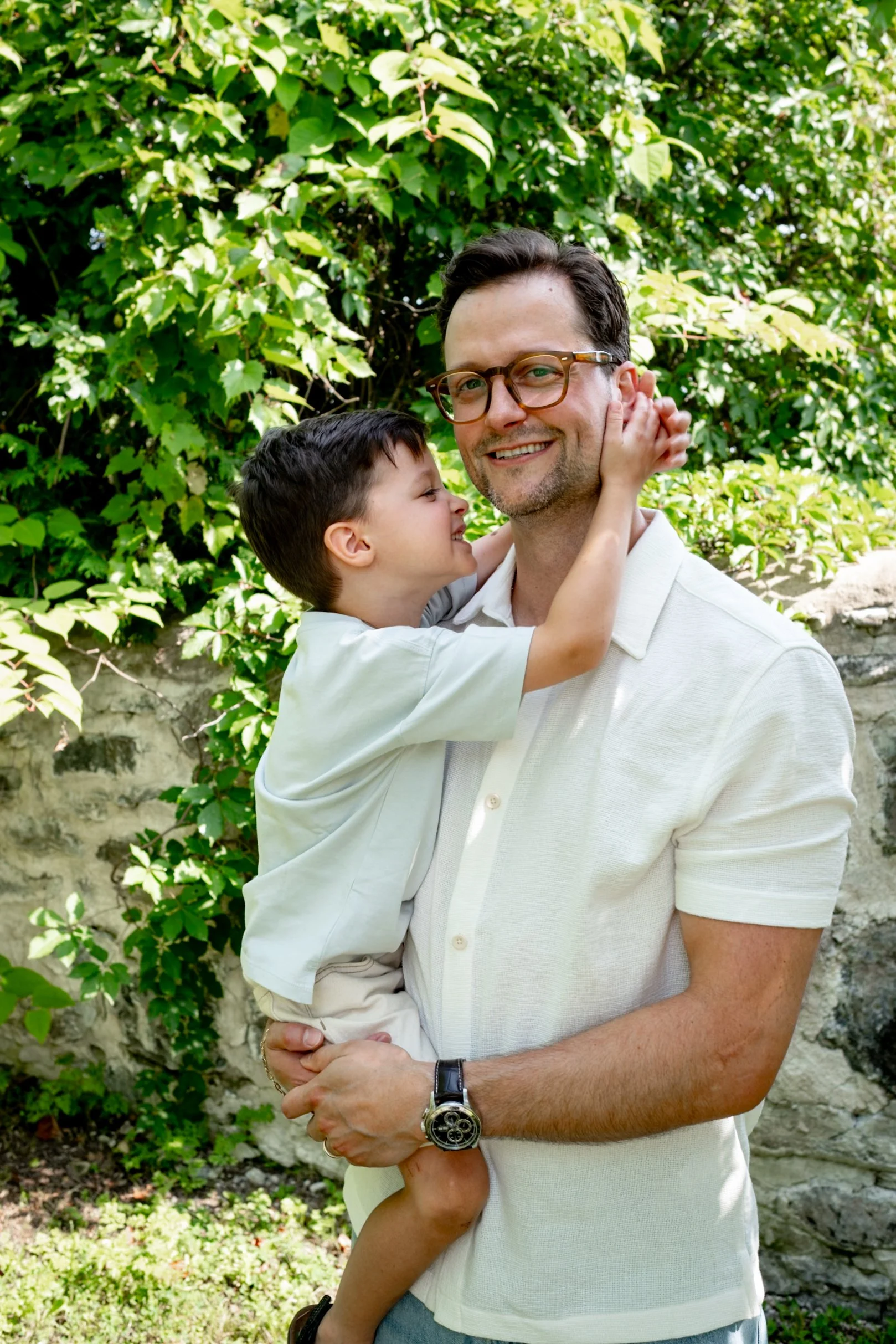 A man holding a young boy outdoors in front of green leafy bushes. The boy is hugging the man and both are smiling.