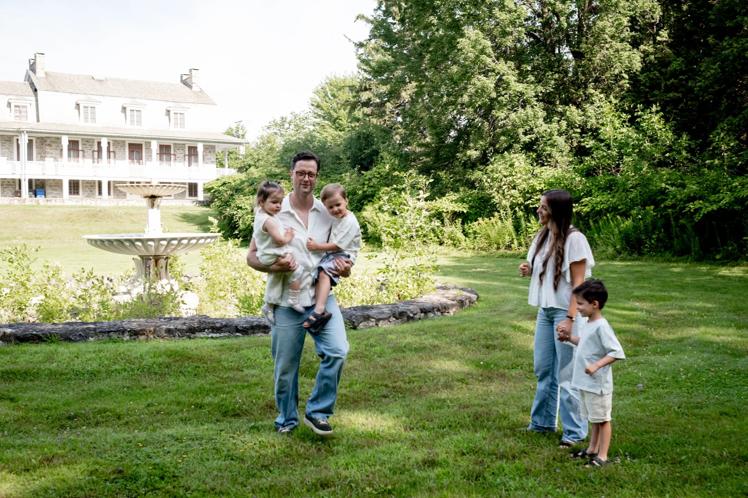 A man holding two children, a woman and two boys standing together on a lawn in front of a large gray house surrounded by trees.