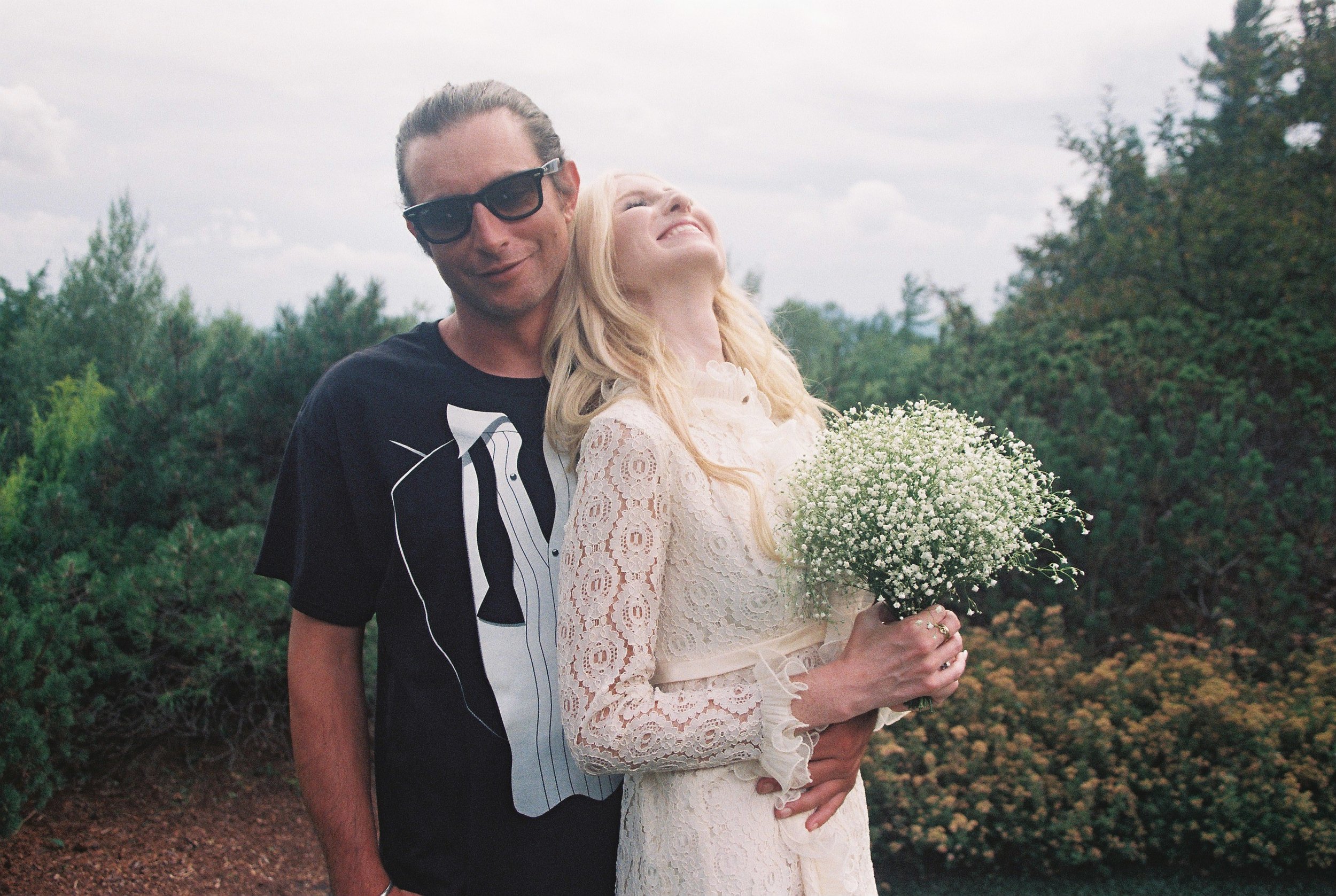 A man and woman stand outdoors, the bride in a white lace dress holding a bouquet of baby's breath flowers, both smiling with her head tilted back and the man smiling, wearing sunglasses, with lush greenery in the background. Outdoor wedding.