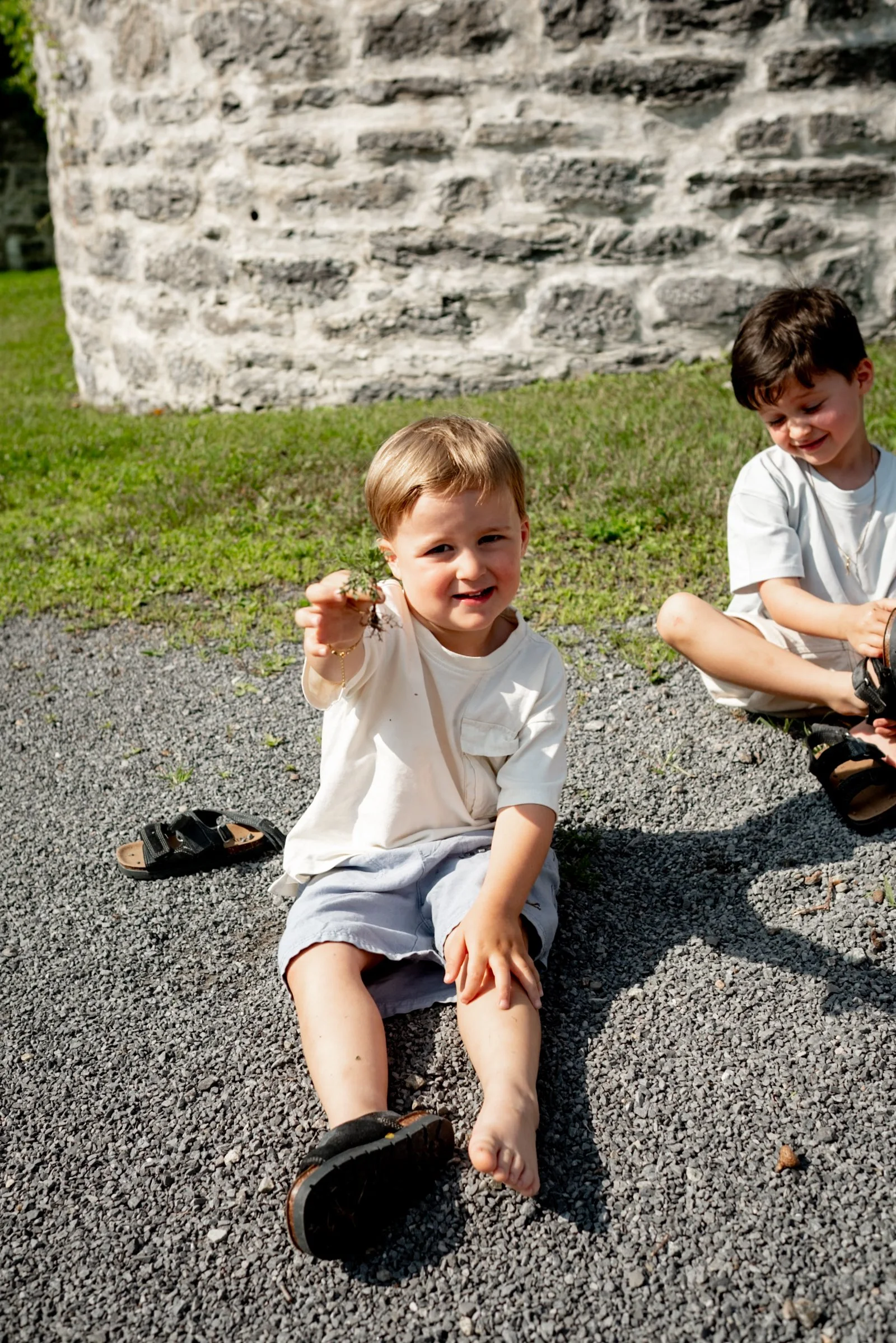 Two young boys sitting on gravel outdoors near a stone building, one holding a small tree branch, smiling and looking at the camera.