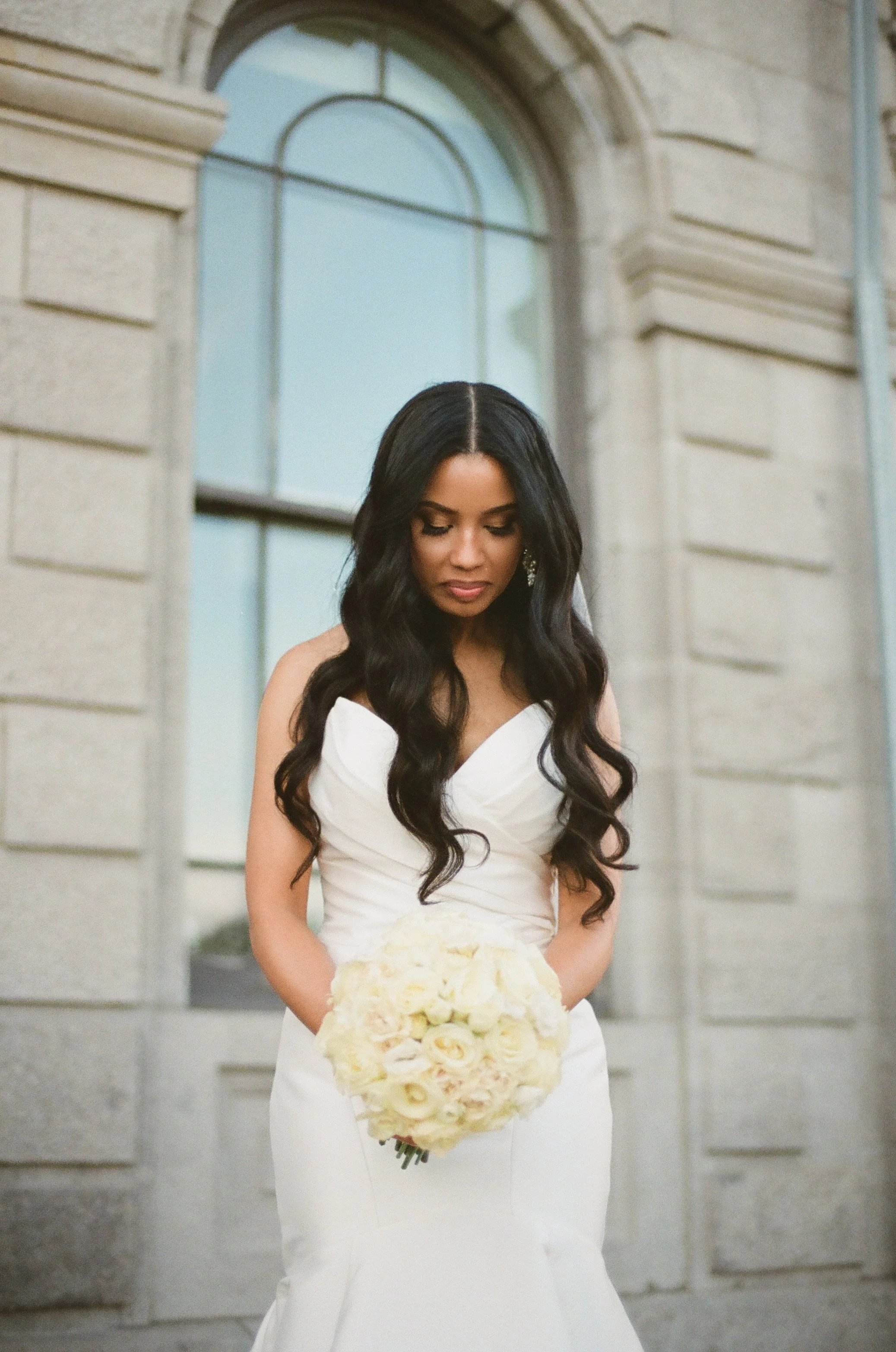 A bride in a white wedding gown holding a bouquet of white roses stands against a stone building with tall arched windows.