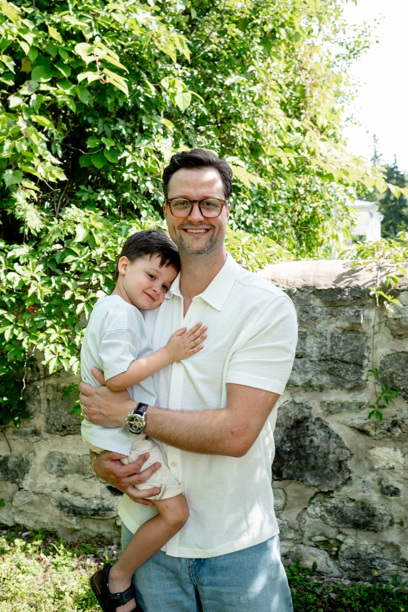 A man holding a young boy in his arms outdoors, surrounded by green bushes and a stone wall in the background, smiling at the camera.