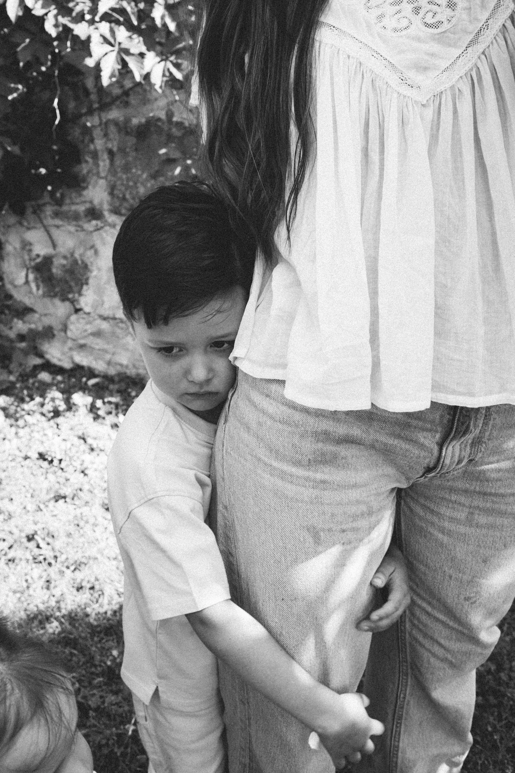 A young boy hugging a woman, possibly his mother, with part of his arm and forehead visible. The woman is wearing jeans and a loose-fitting shirt. The background shows outdoor plants or foliage.
