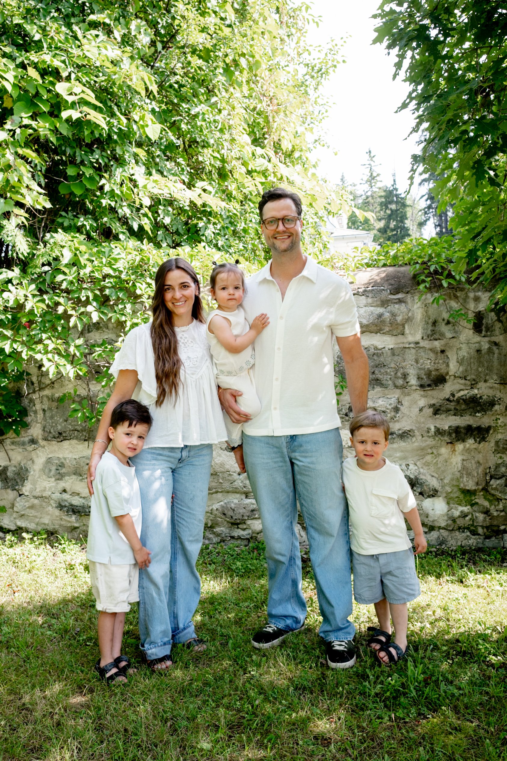 A family of five, including two children, stands outdoors in front of a stone wall and lush green trees, smiling at the camera.