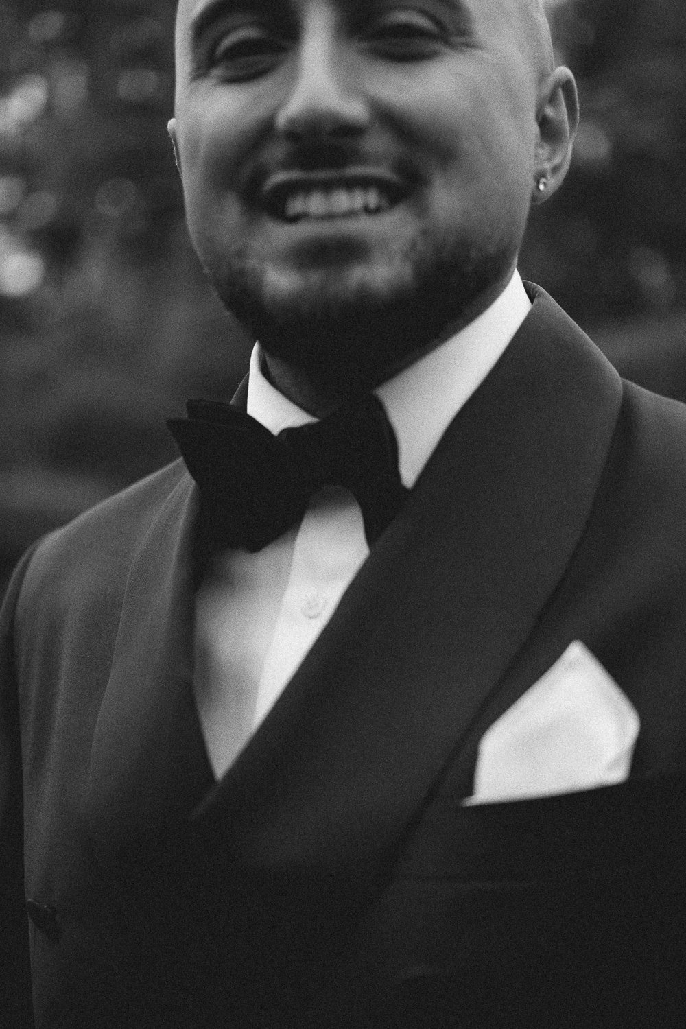 Handsome groom dressed in a tuxedo with a bow tie, white shirt, and pocket square, outdoors in black and white.