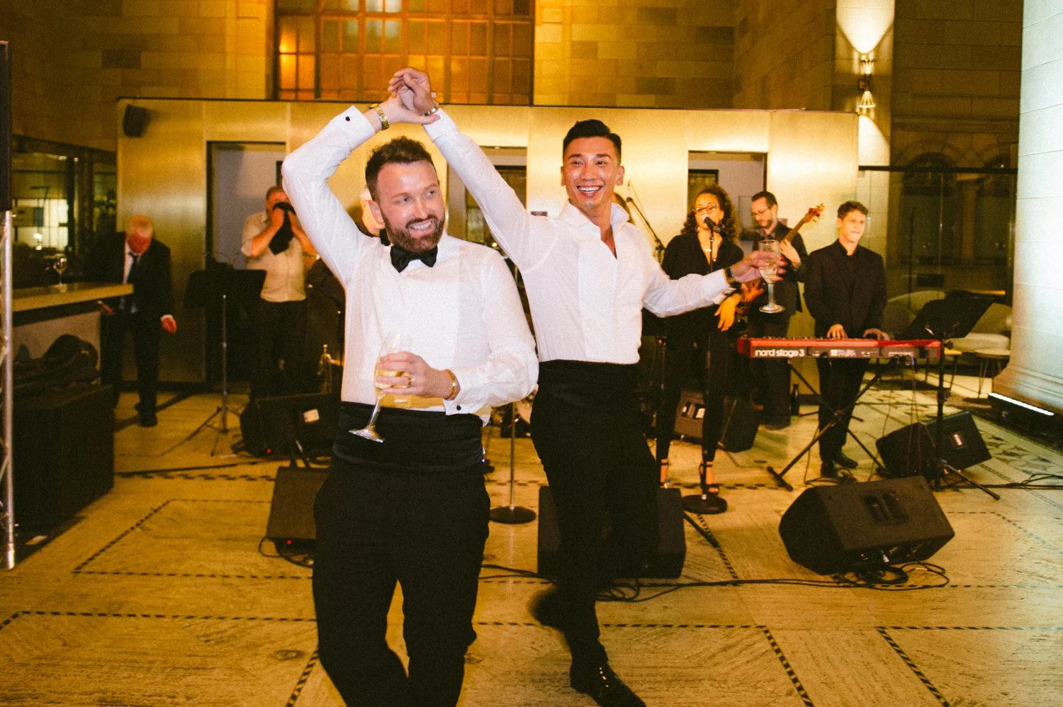 Two men dancing at a party with a band playing in the background. The man on the left is wearing a white shirt and a black bow tie, holding a drink, and the man on the right is in a white shirt and black pants, smiling and holding a glass. The band m