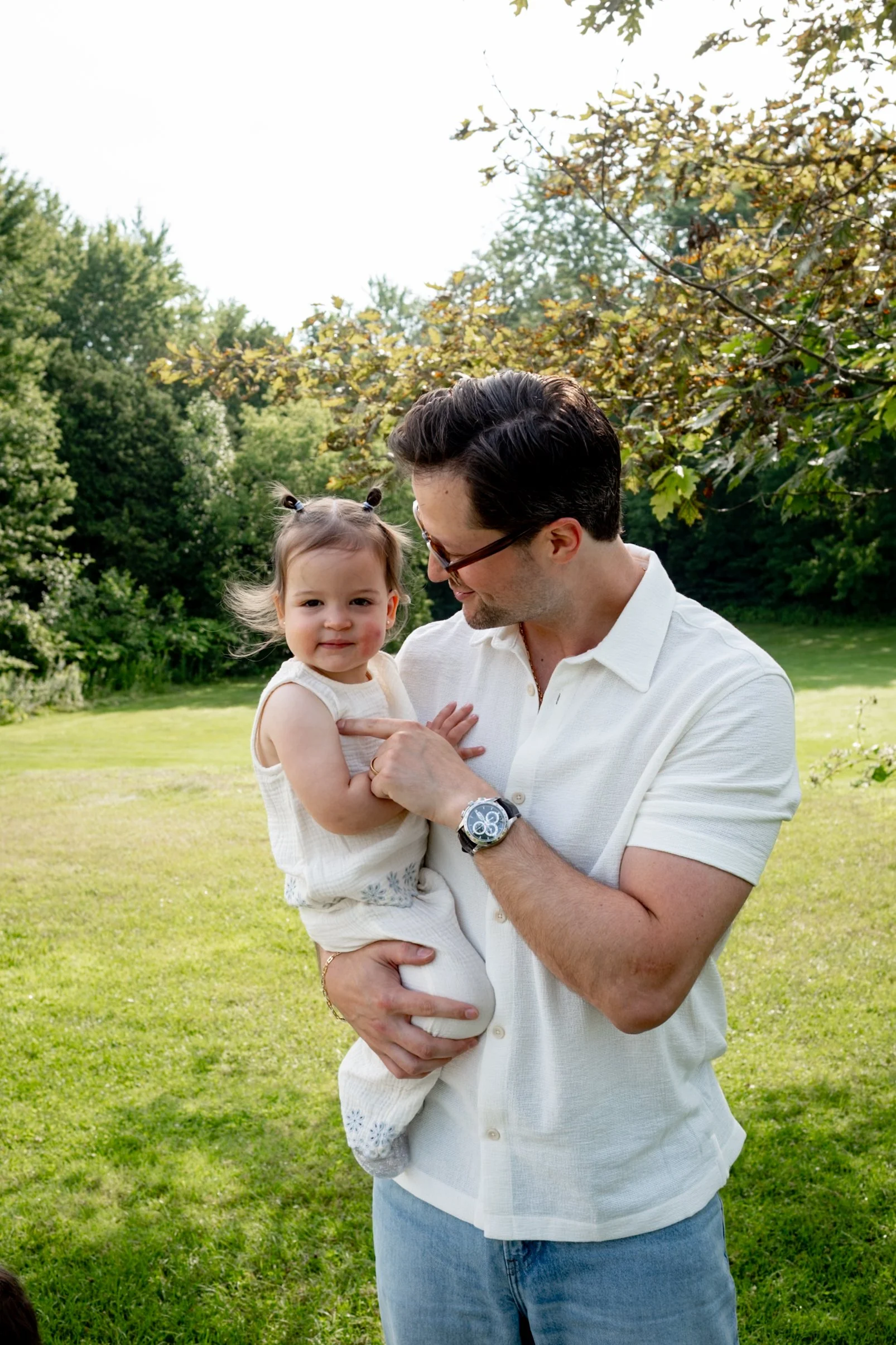A man holding a young girl in an outdoor park with trees and green grass, smiling and looking at her.