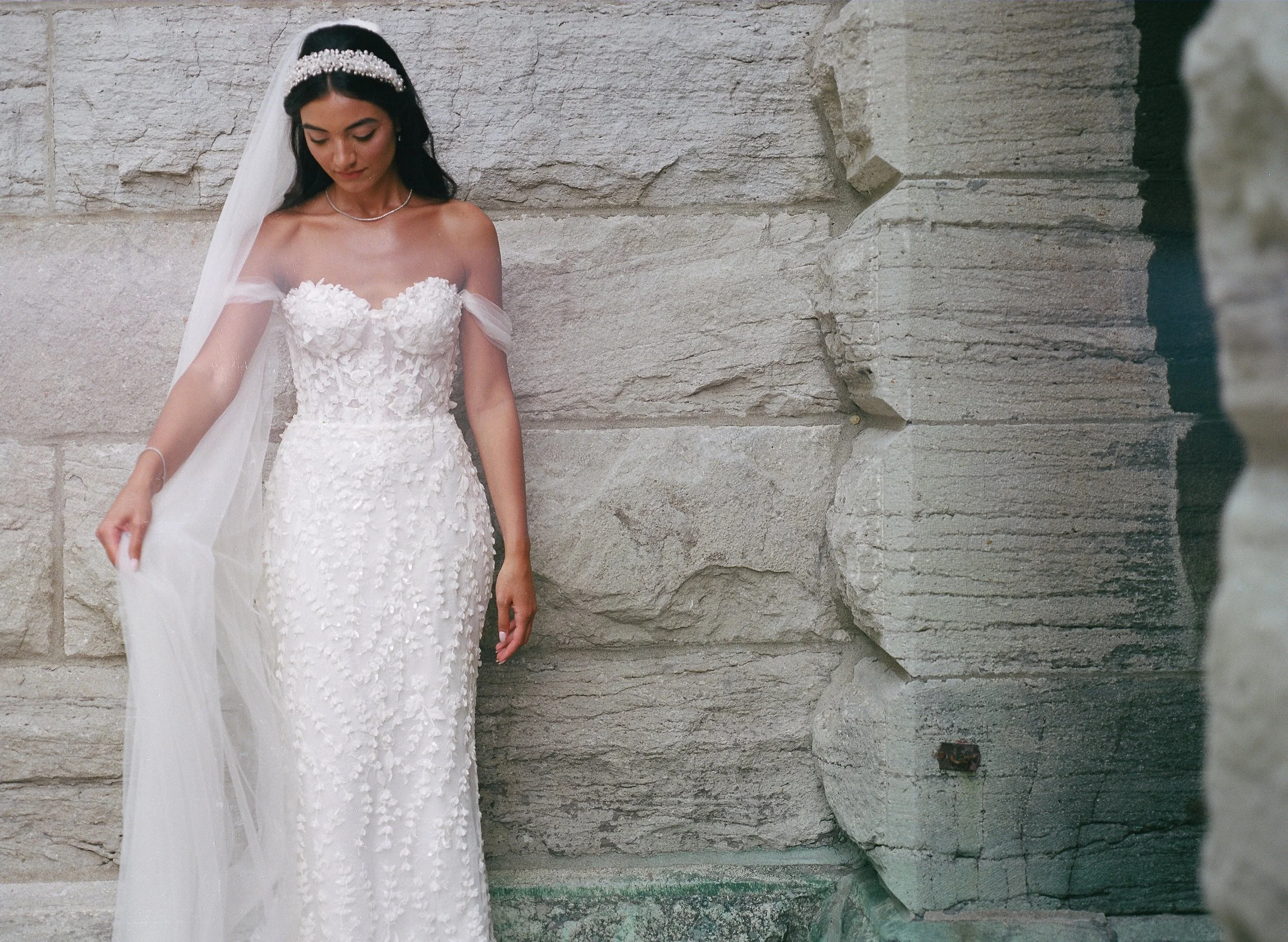 A bride wearing a white wedding dress with floral embellishments, standing against a stone wall, holding a sheer veil in one hand, with dark hair and a pearl headband.