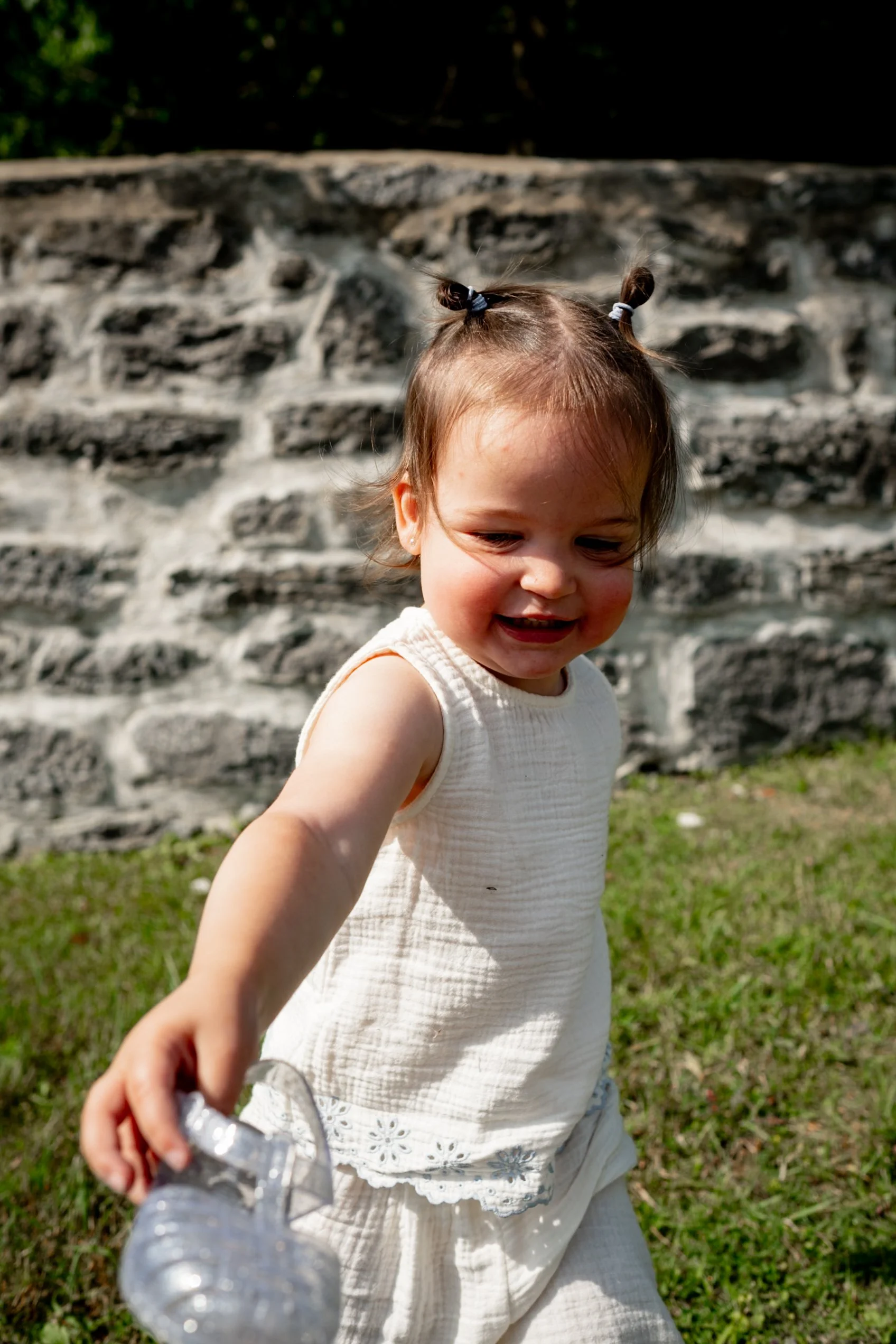 A young girl with brown hair styled in two small buns, wearing a white sleeveless dress with embroidery, playing outside near a stone wall with greenery in the background.