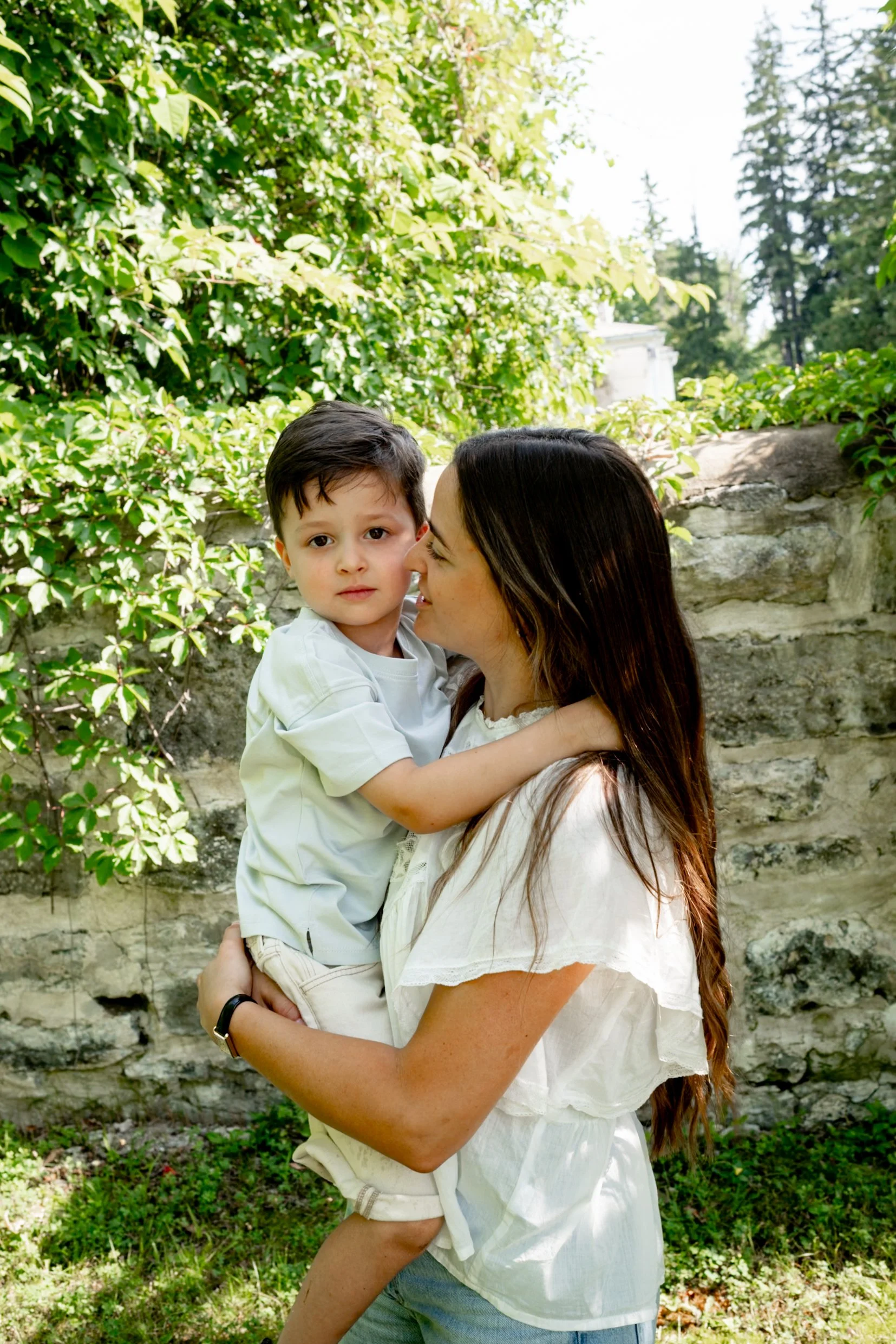 A woman holding a young boy in an outdoor setting with green bushes and a stone wall in the background.